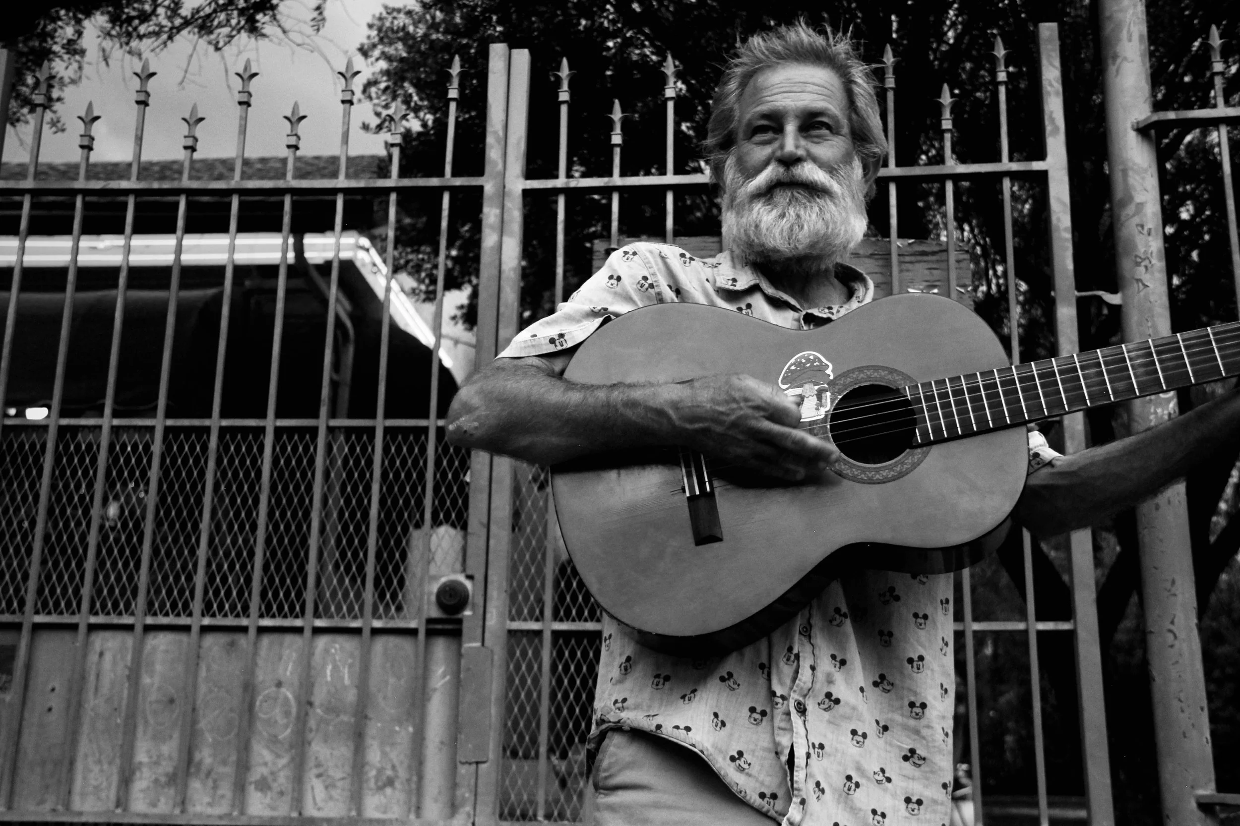 A man with a gray beard and, wearing a shirt with Mickey Mouse prints, stands outdoors in front of a metal fence while playing an acoustic guitar.