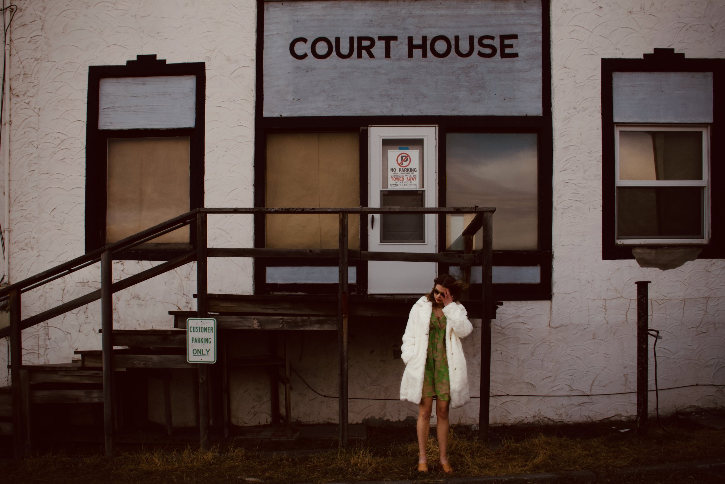 A woman in a white coat and green dress standing outside a building with a sign that reads "Court House". The building has two large windows and a door with a "No Parking" sign. There are steps leading up to the entrance and a small sign indicating c
