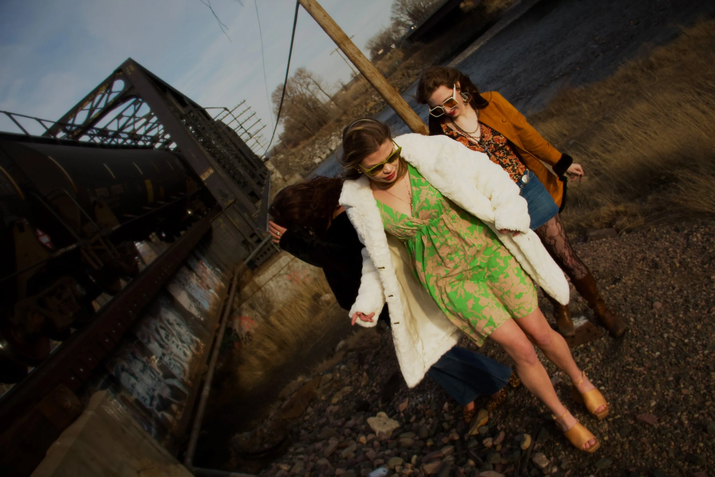 Three women dressed in vintage 1970s-style clothing standing outdoors by train tracks, with trees and utility poles in the background, during late afternoon or early evening.