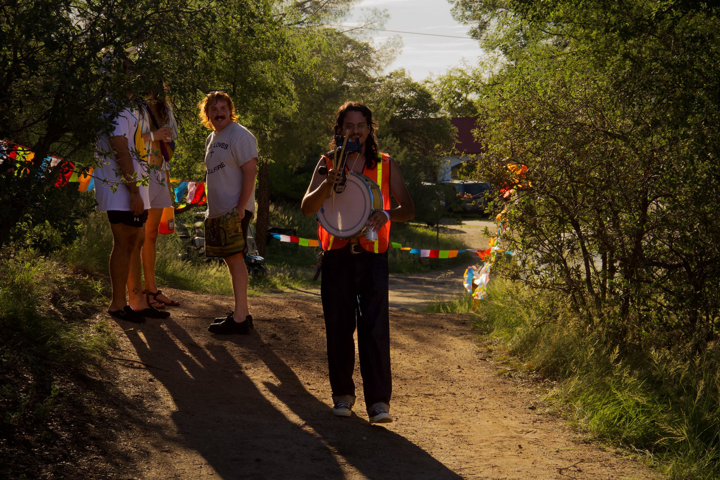 Group of people walking along a dirt path surrounded by trees, with colorful decorations hanging between the trees and sunlight filtering through.