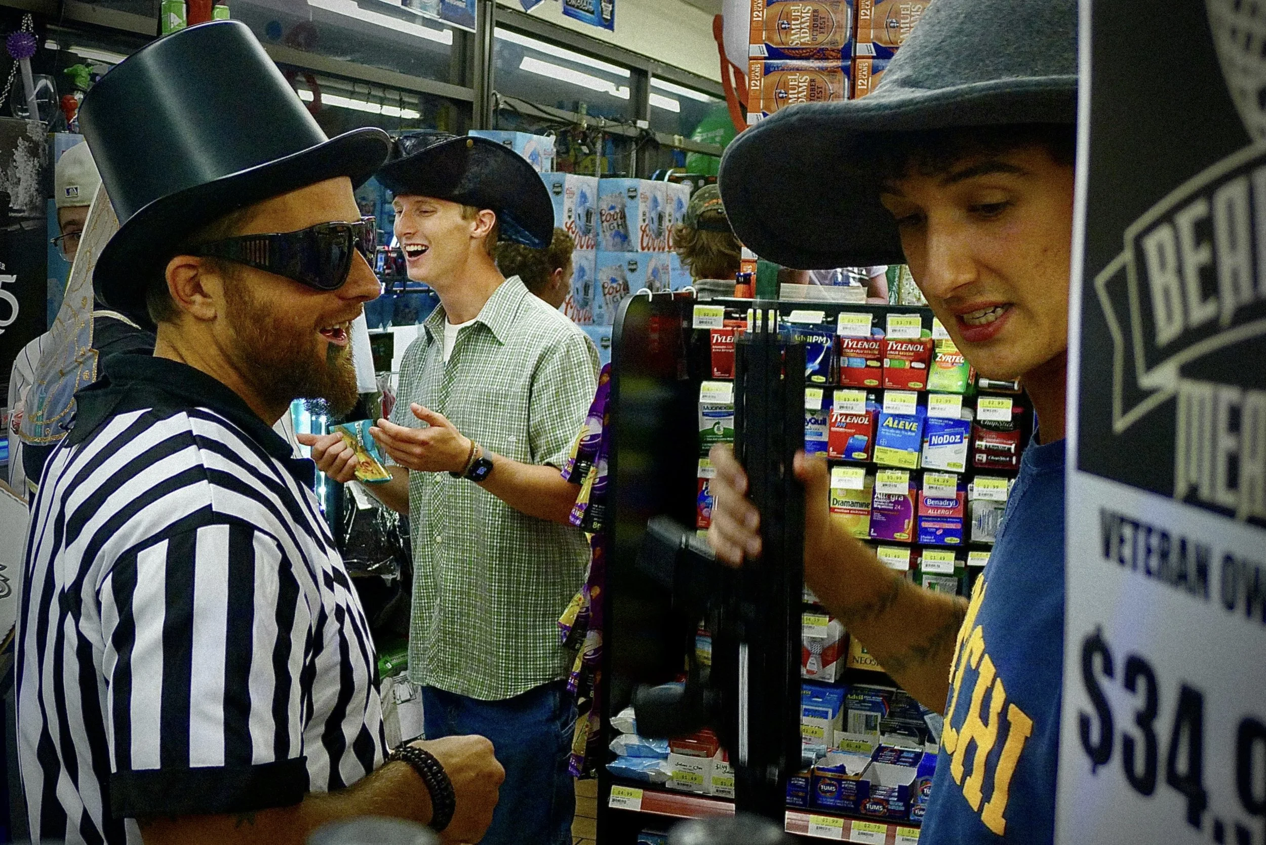 People shopping at a pharmacy, wearing costumes and masks, including a man with a striped referee shirt, a woman with a witch or wizard hat, and a person wearing a large black hat and sunglasses, surrounded by shelves of medicines and healthcare prod