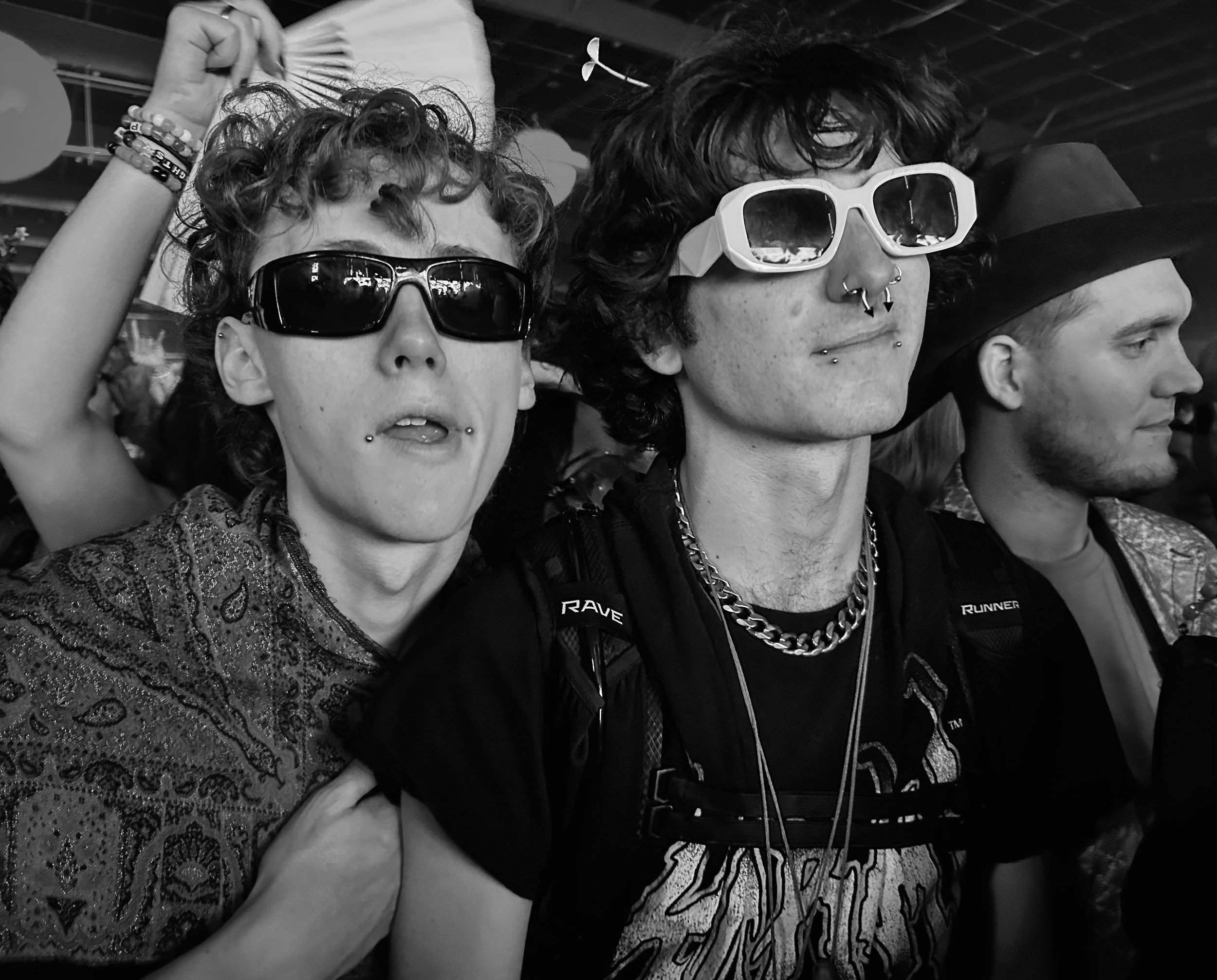 Three individuals wearing sunglasses, with two of them showing vulnerability with facial expressions, and a woman in the foreground with her mouth open and tongue slightly out. They are close together, with a dark indoor or outdoor setting in the bac