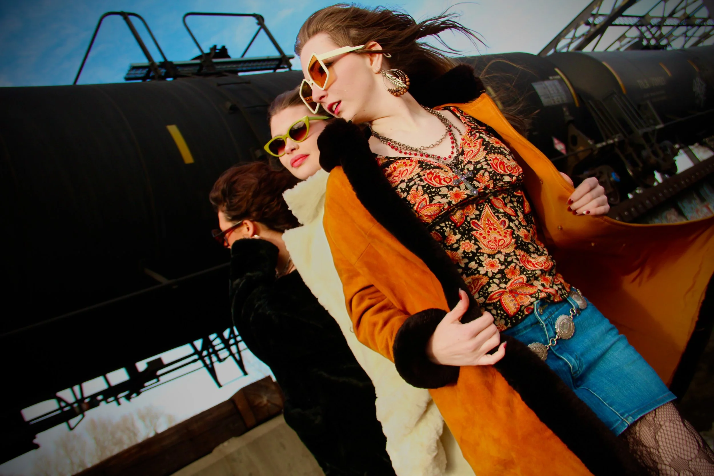 Three women standing close together, wearing colorful outfits, sunglasses, and jewelry, outdoors with an industrial structure in the background.