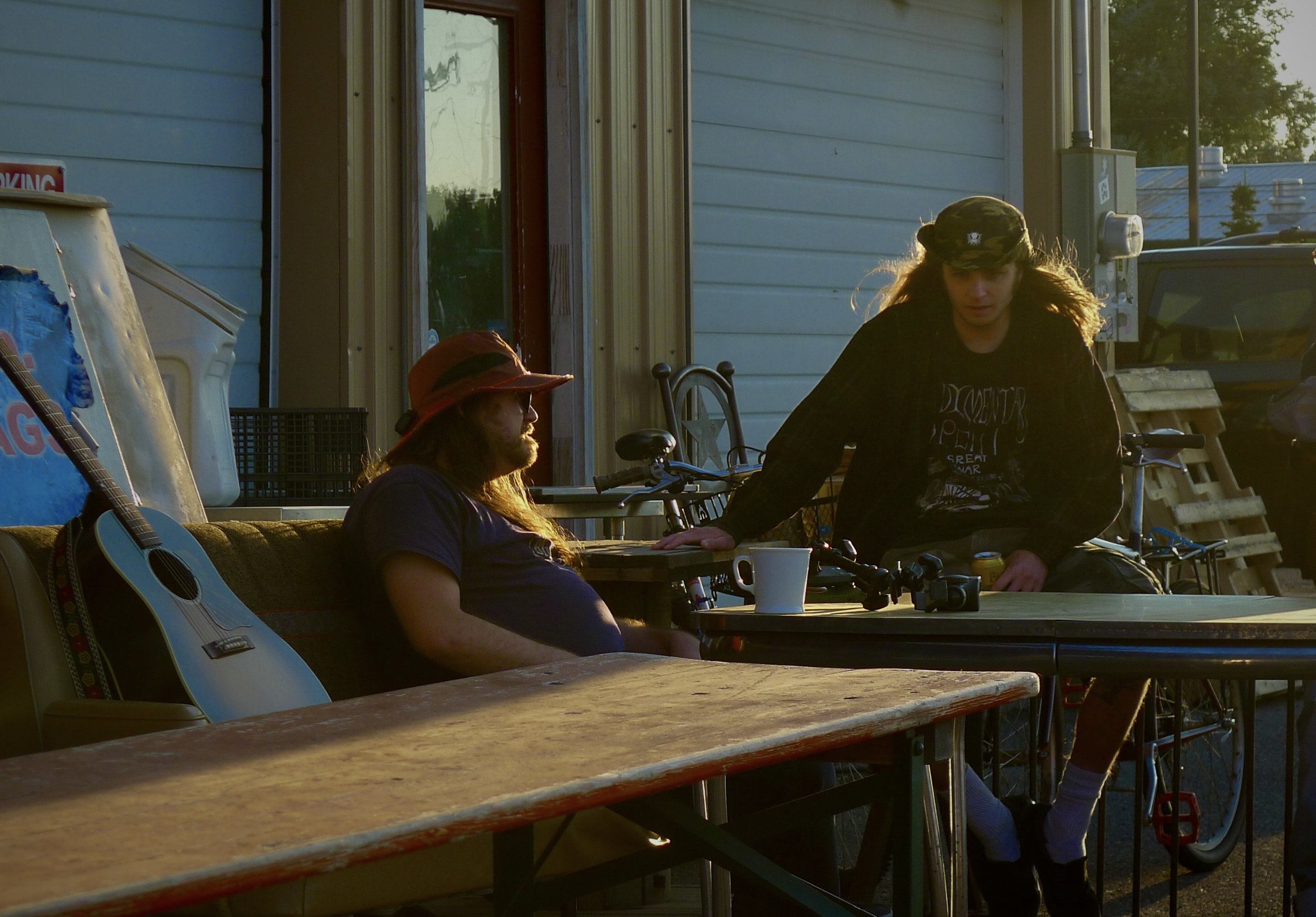 Two women sitting at a table outside, one with long hair wearing a sunhat, the other with long hair wearing a camouflage cap, with bicycles near them and a guitar leaning against a chair.