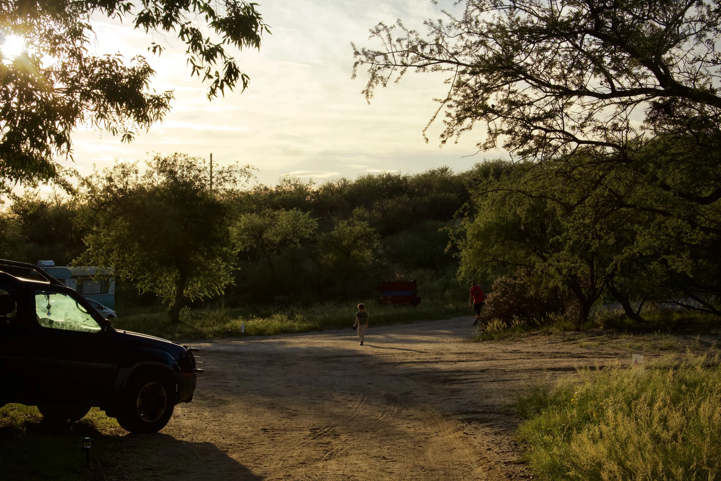 A dirt parking lot surrounded by trees with the sun setting in the background, casting a warm glow. Two children walking in the distance, with cars parked on the left side.