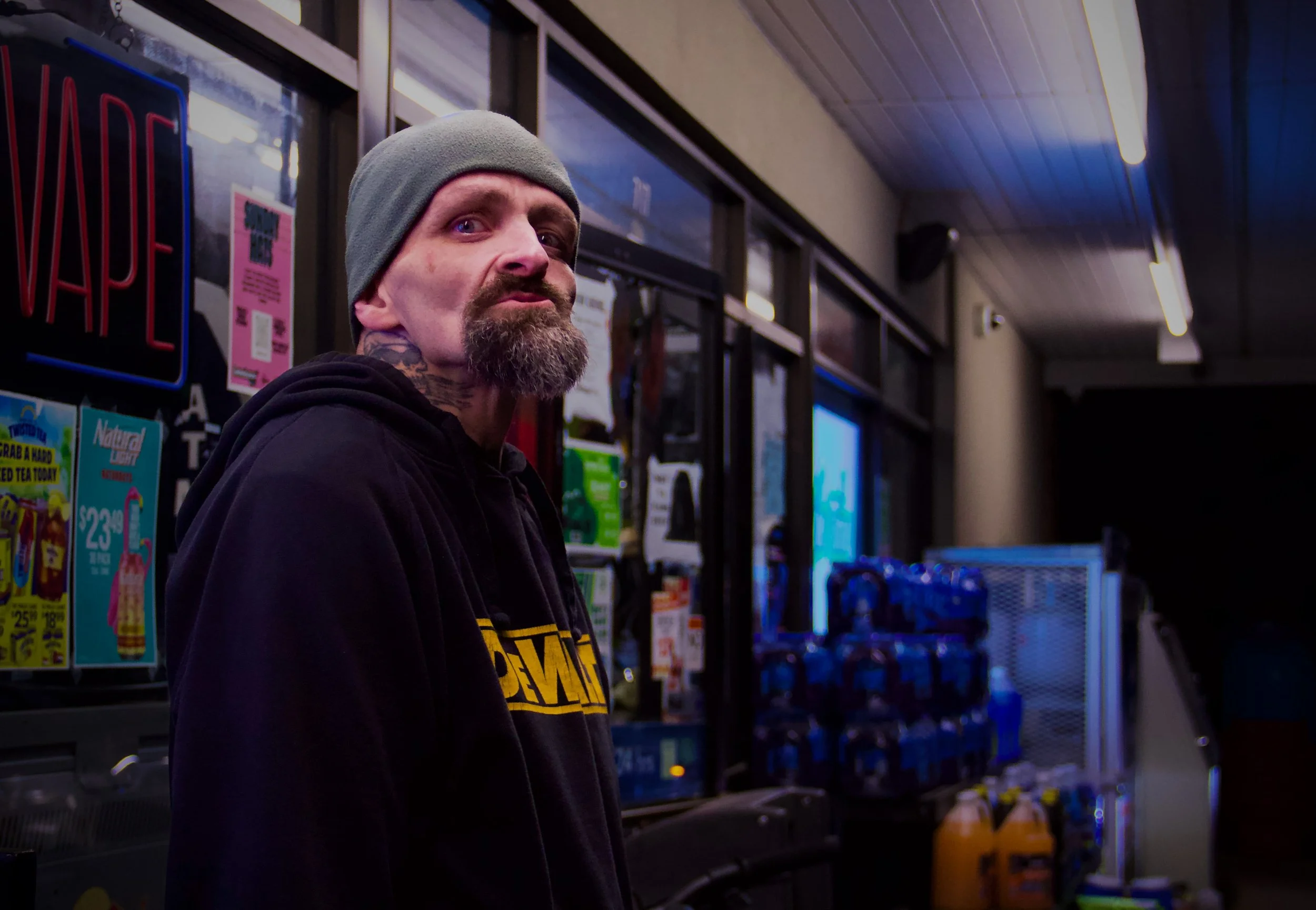 A man with a gray beanie and a beard looks at the camera inside a store. Shelves behind him are stocked with various products, including snacks and drinks.