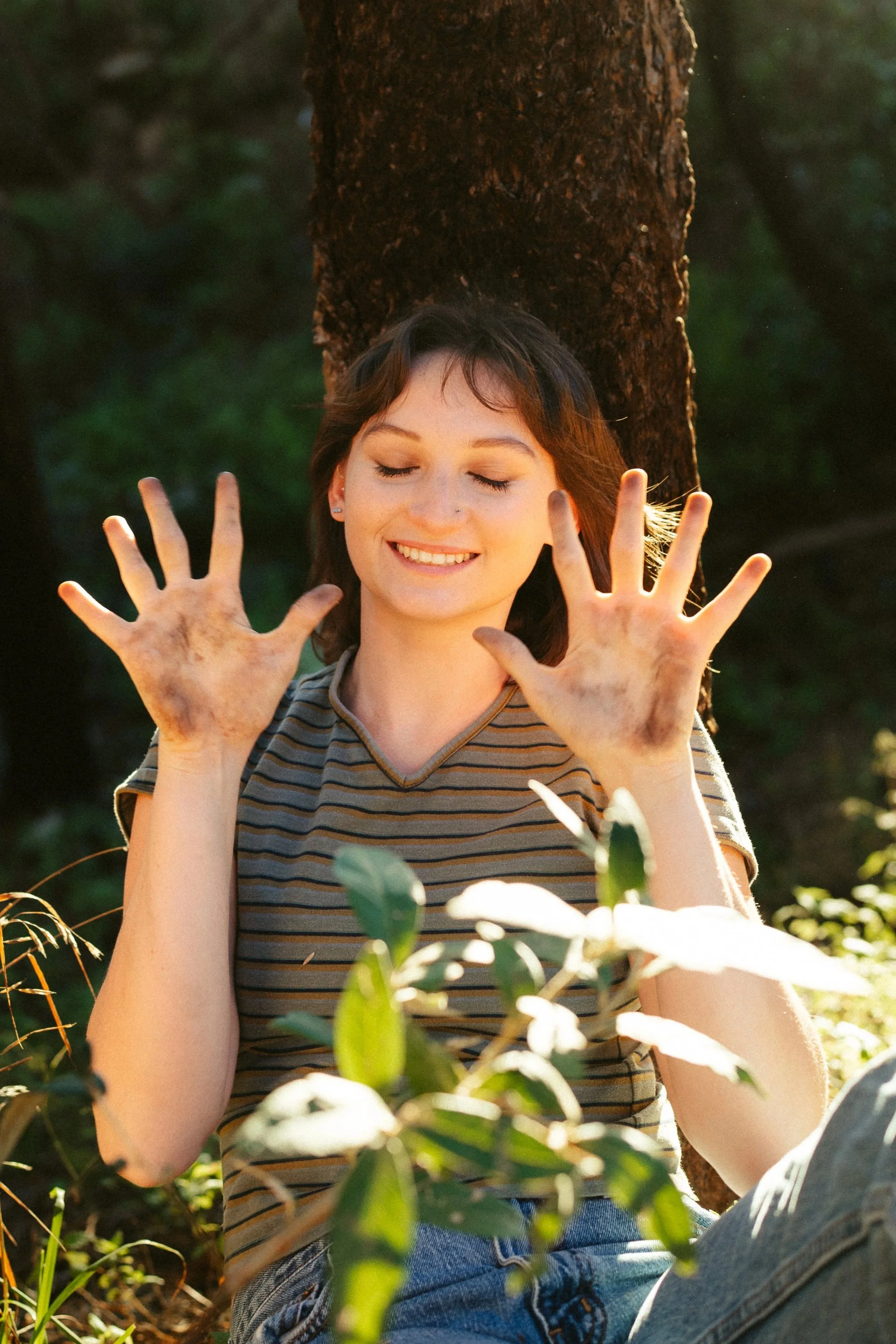 A young woman with short brown hair and light skin sitting outdoors in a natural setting, smiling with her eyes closed, wearing a striped t-shirt and jeans, with her hands raised and dirt on them.