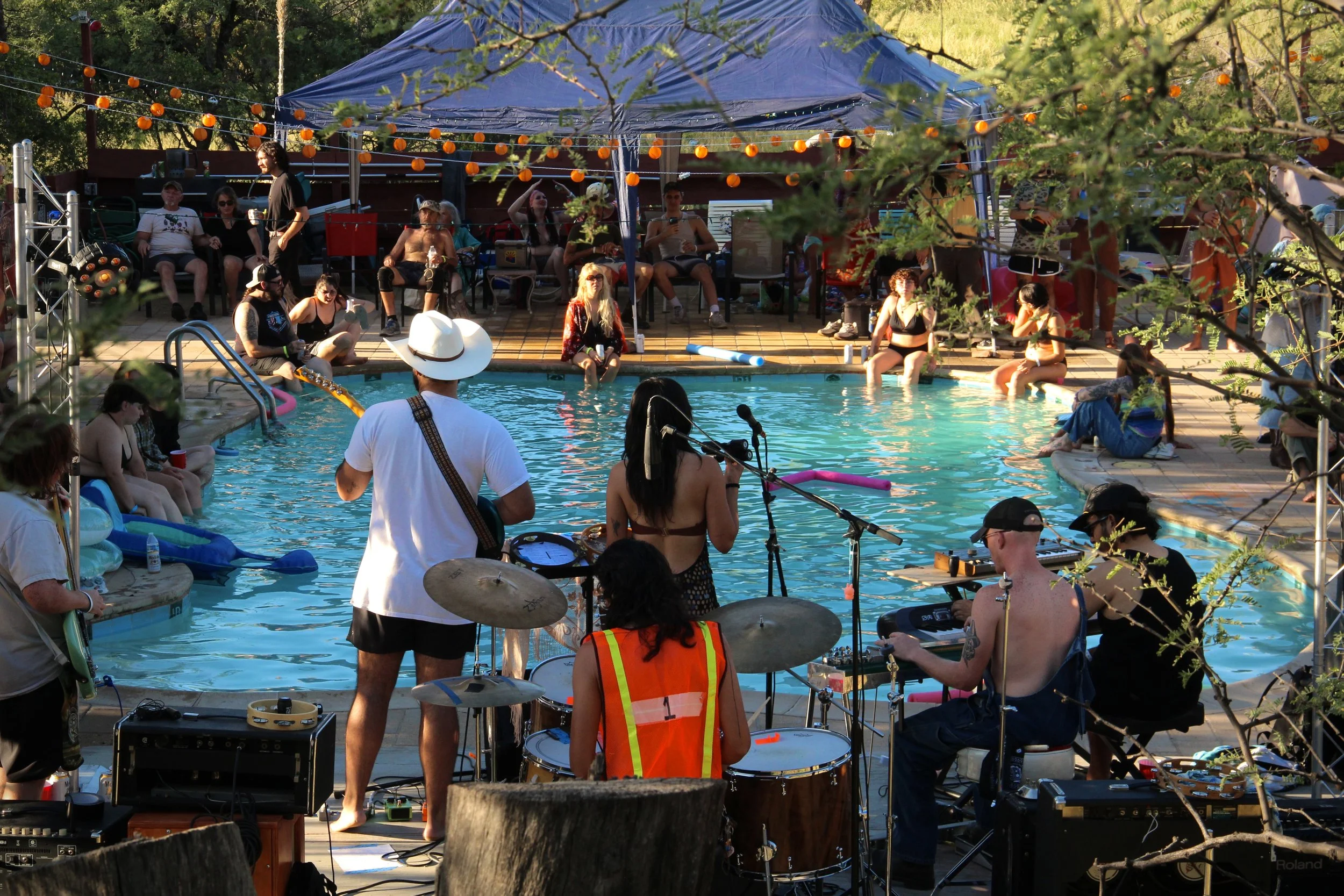 People gathered around a swimming pool enjoying a party, with a band performing music in the foreground and past the pool, trees and string lights in the background.