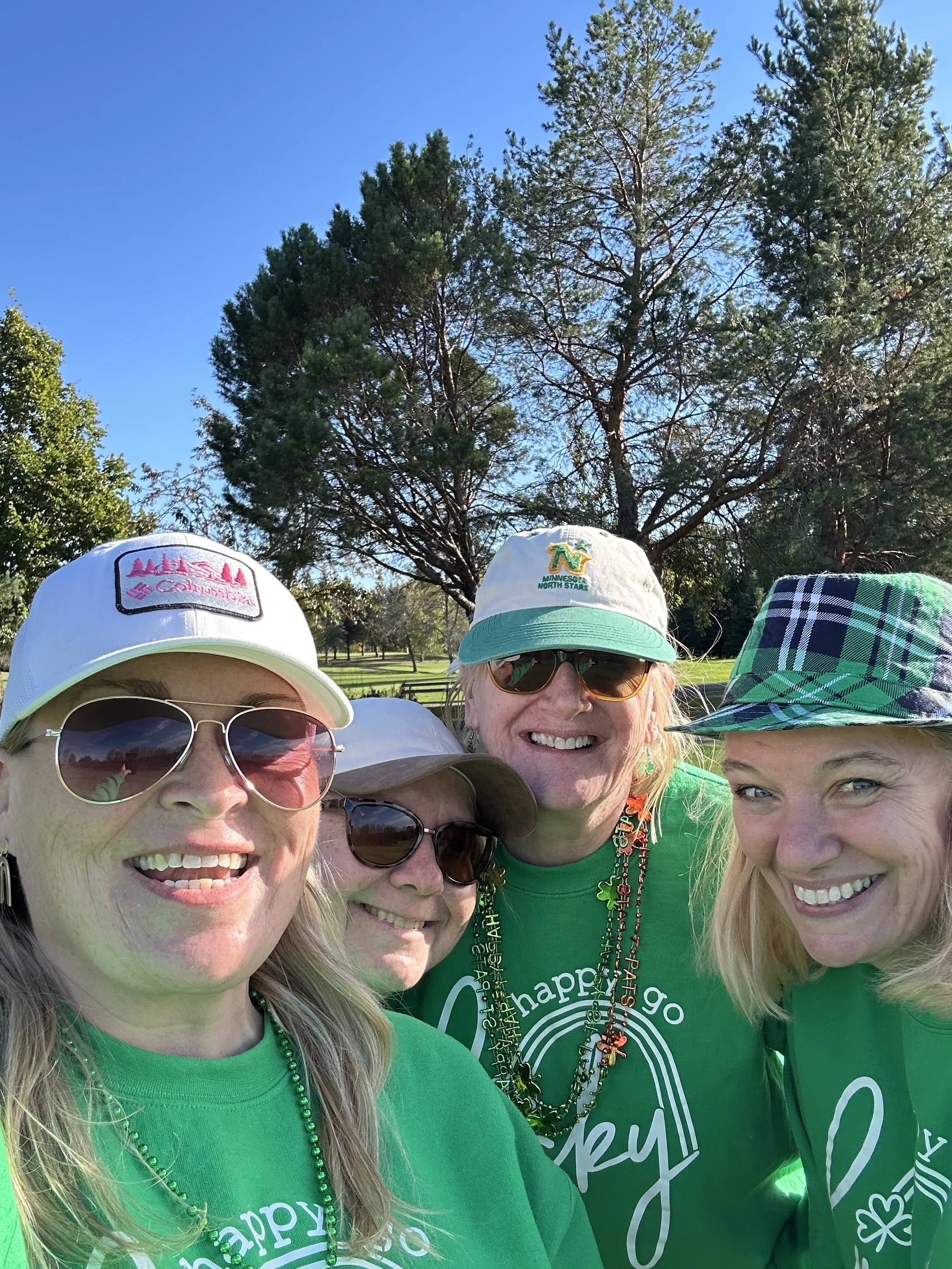 Group of women celebrating, wearing green shirts and St. Patrick's Day accessories, outdoors in a park with tall trees and blue sky in the background.