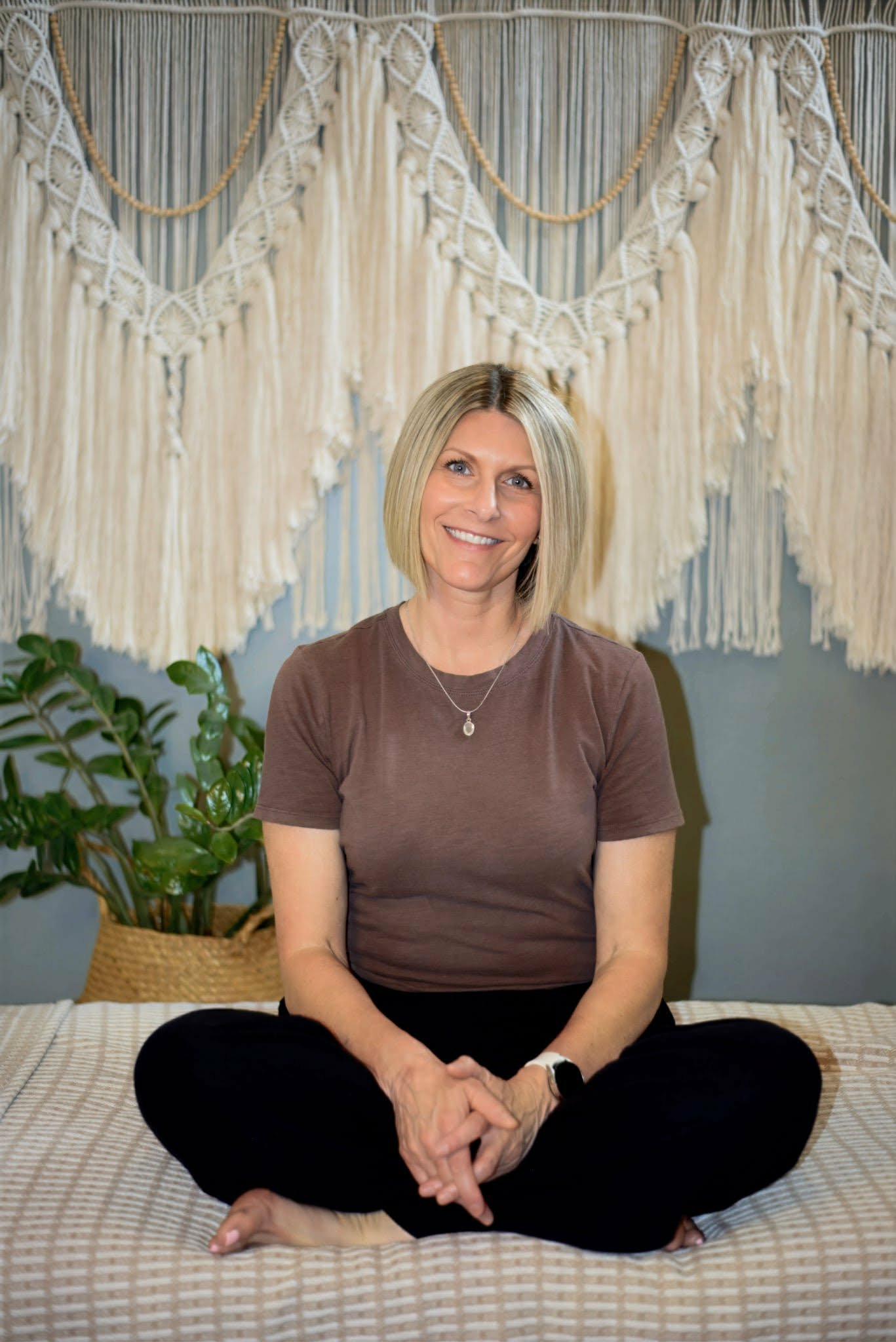 Jessica, a licensed massage therapist, sitting cross-legged on a massage table with a checkered blanket, smiling, with green houseplants and a decorative macramé wall hanging in the background.