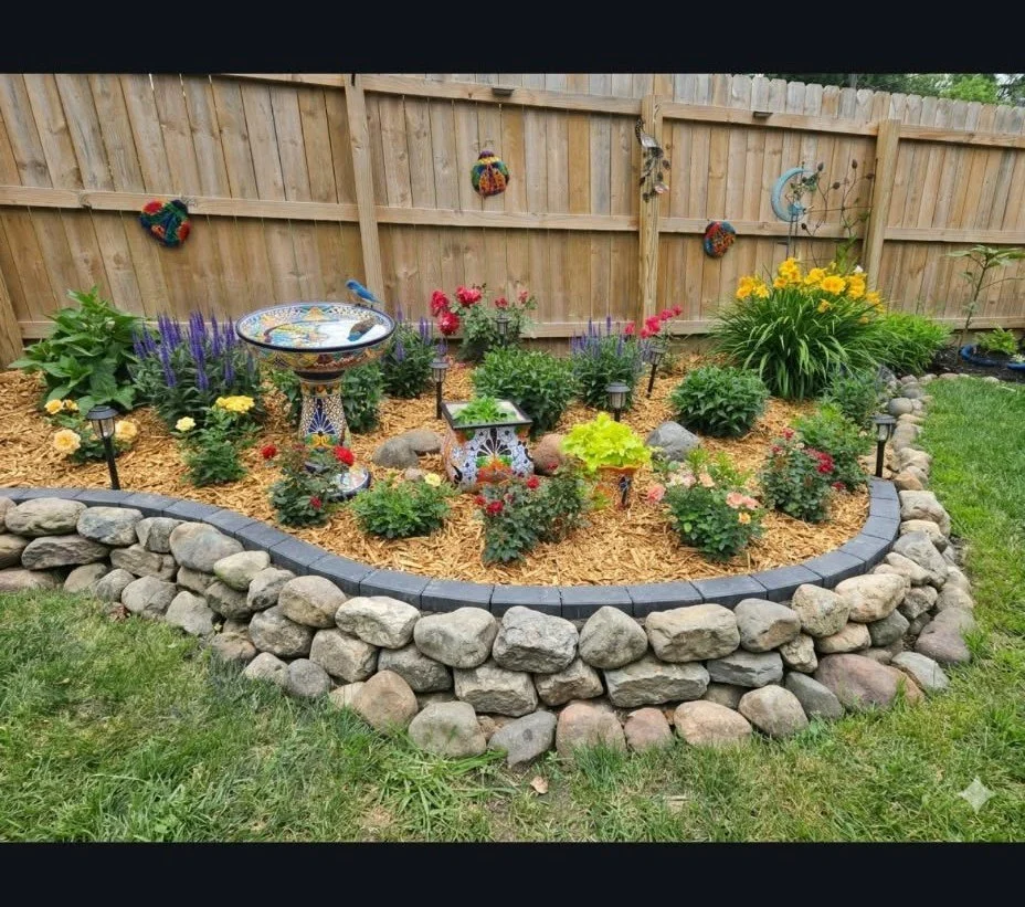 A colorful garden bed with various flowers, decorative statues, and solar lights, surrounded by a stone border and a wooden fence in the background.