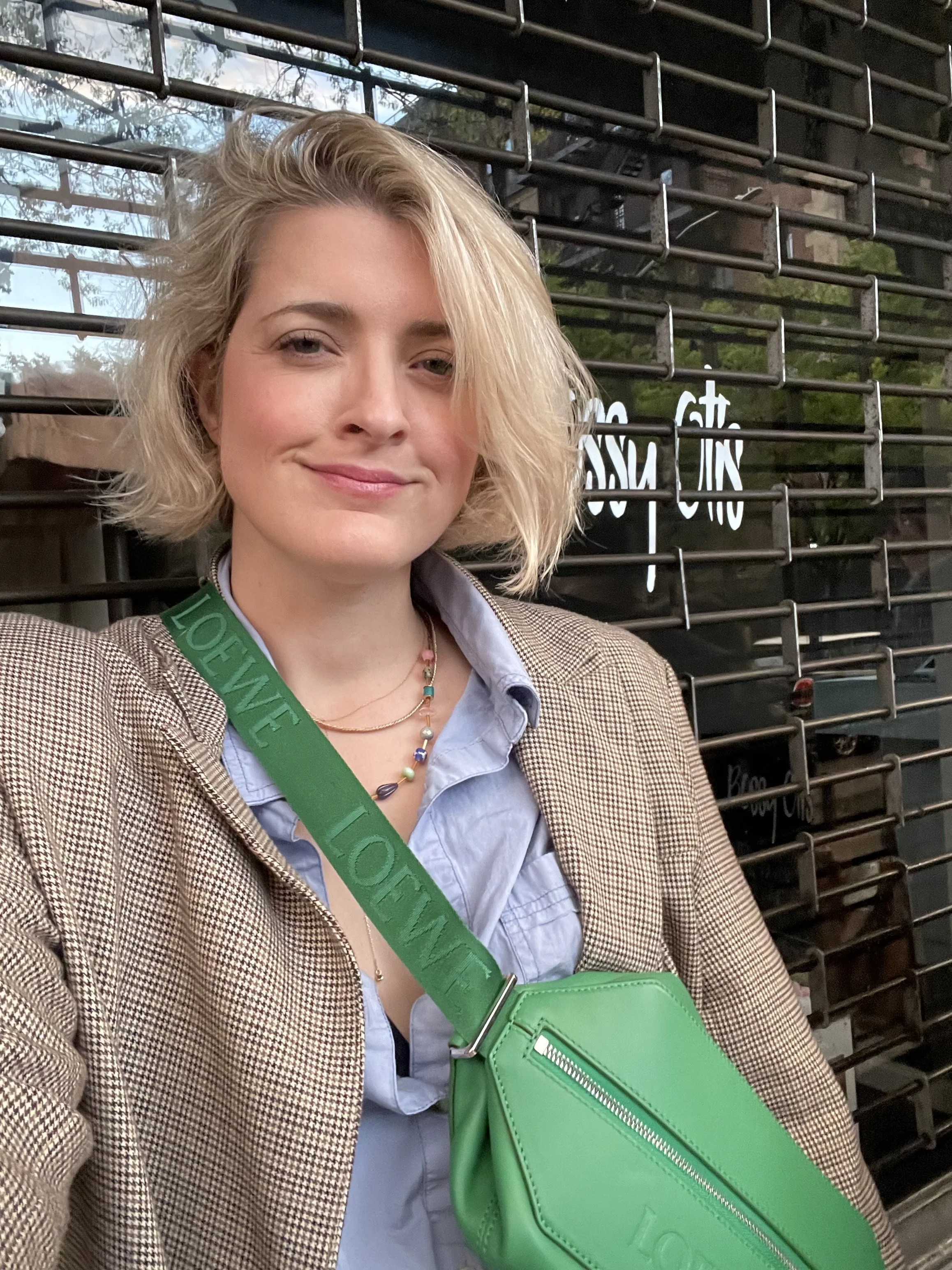 A woman with short blonde hair smiling, wearing a beige plaid blazer, a blue shirt, a colorful beaded necklace, and a green Louis Vuitton crossbody bag. She is standing outdoors in front of a black metal fence with reflective glass panels, trees, and a sign with white letters in the background.
