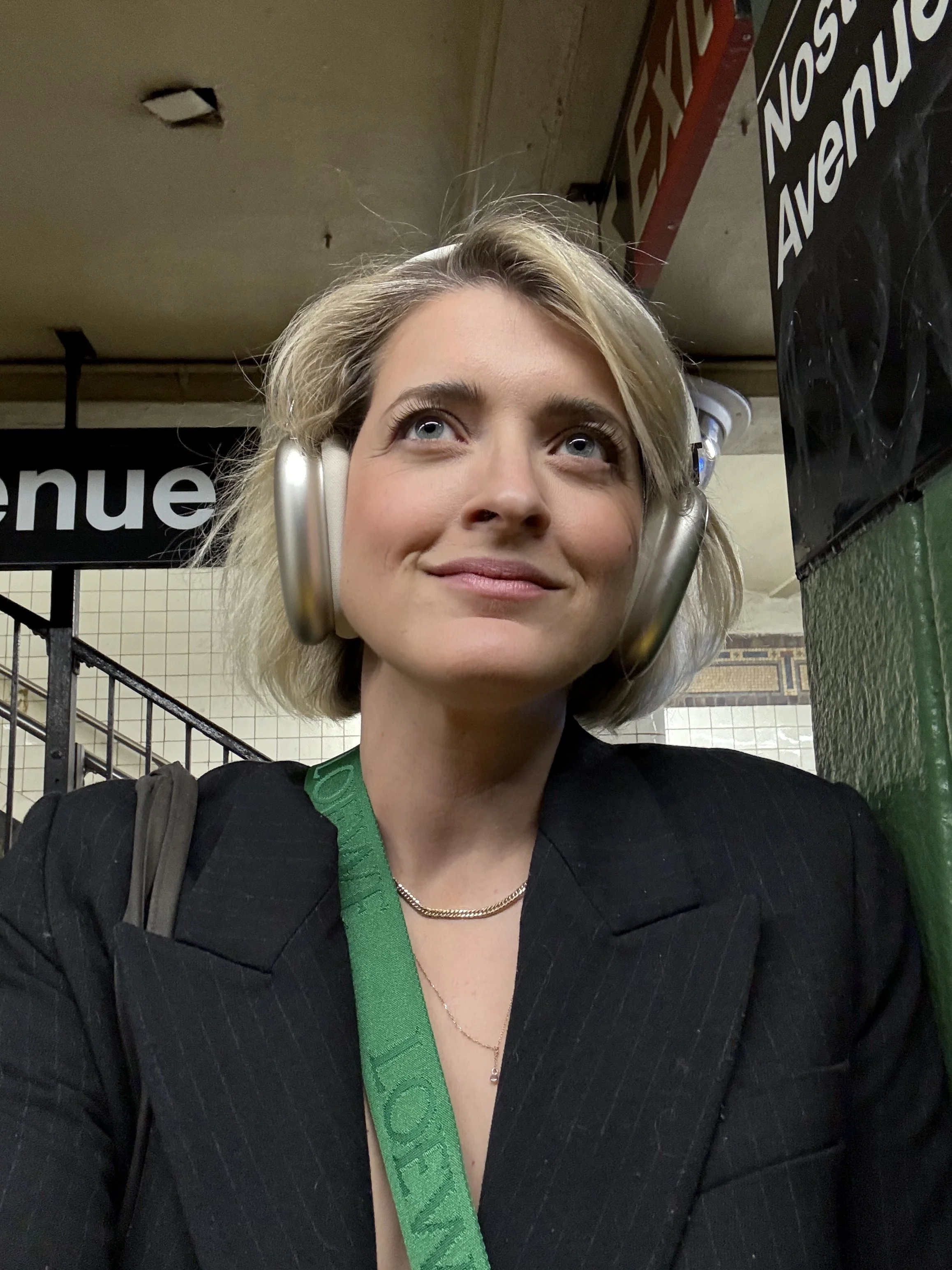Close-up of a woman with blonde hair and light blue eyes wearing headphones, a black pinstripe blazer, a green strap, a gold necklace, and a silver pendant in subway station with tiled walls and signs.