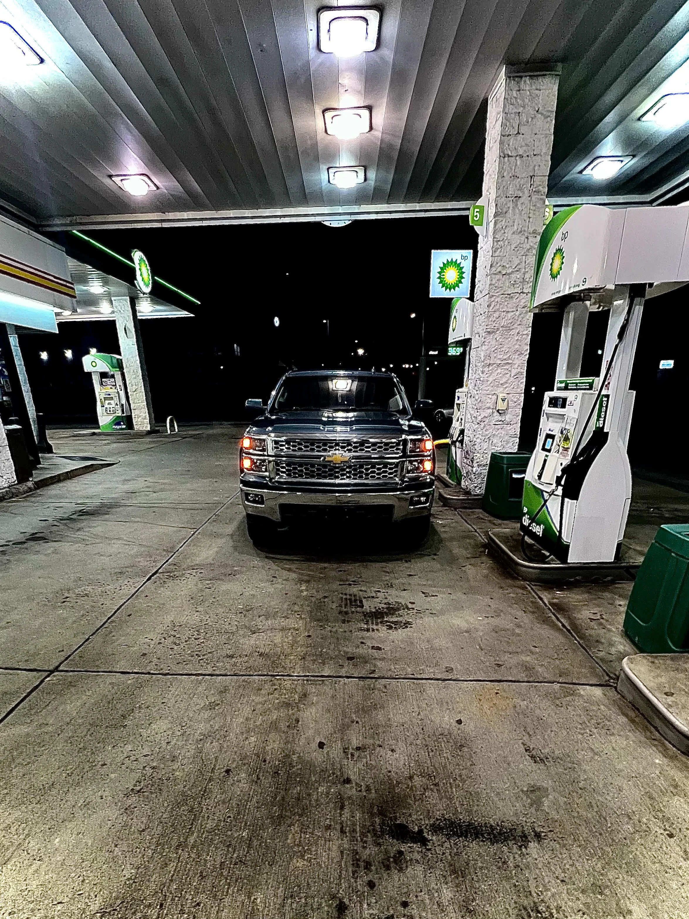 Night scene at a BP gas station with a black Chevrolet Silverado pickup truck parked at the fuel pump station, illuminated by overhead lights, with gas pumps and signs visible on the right side of the image.
