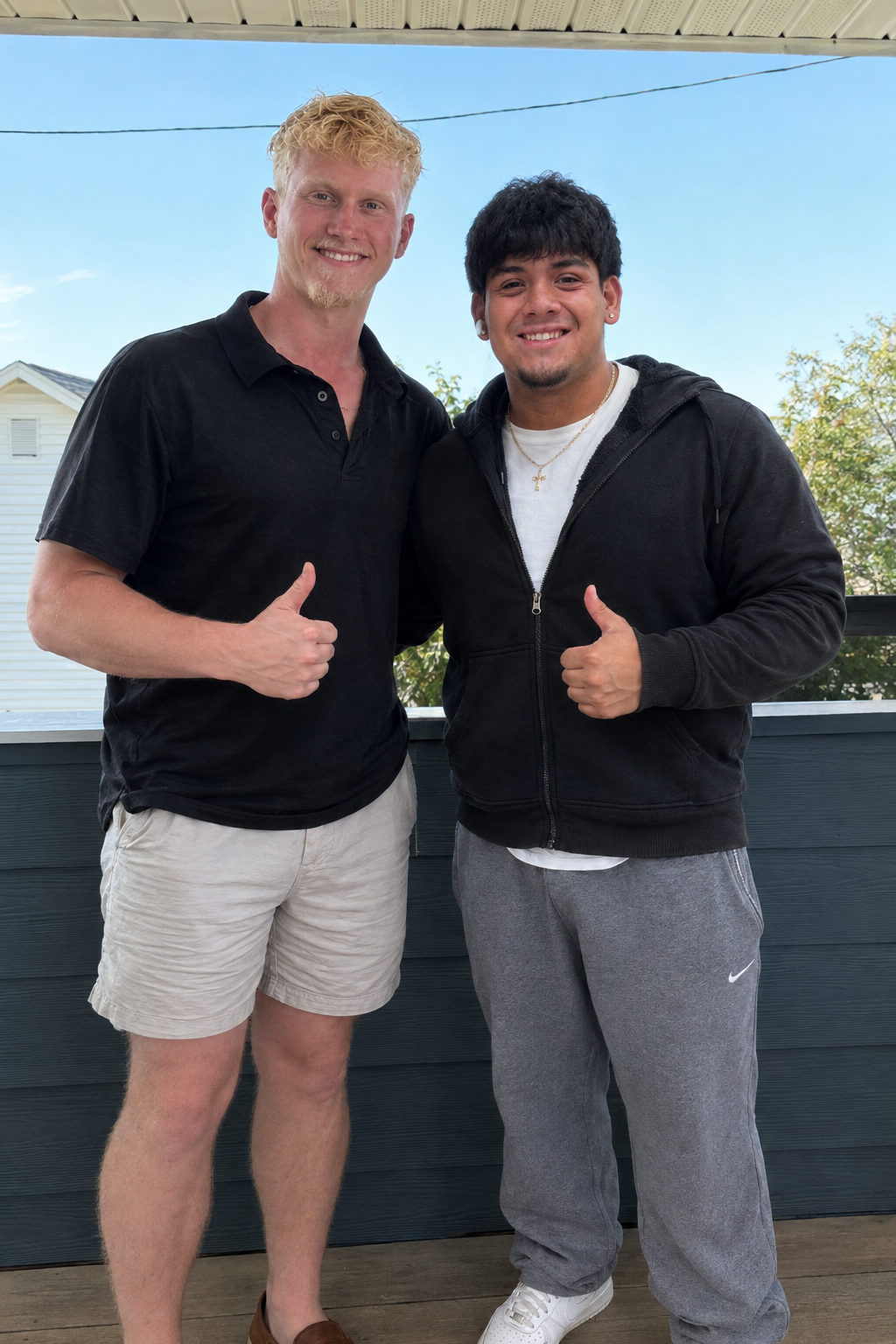 Two young men standing on a porch, smiling and giving thumbs up, with a house and trees in the background.