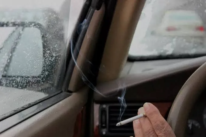 Hand holding a lit cigarette inside a car during rain, with rain-covered windshield and rearview mirror visible.