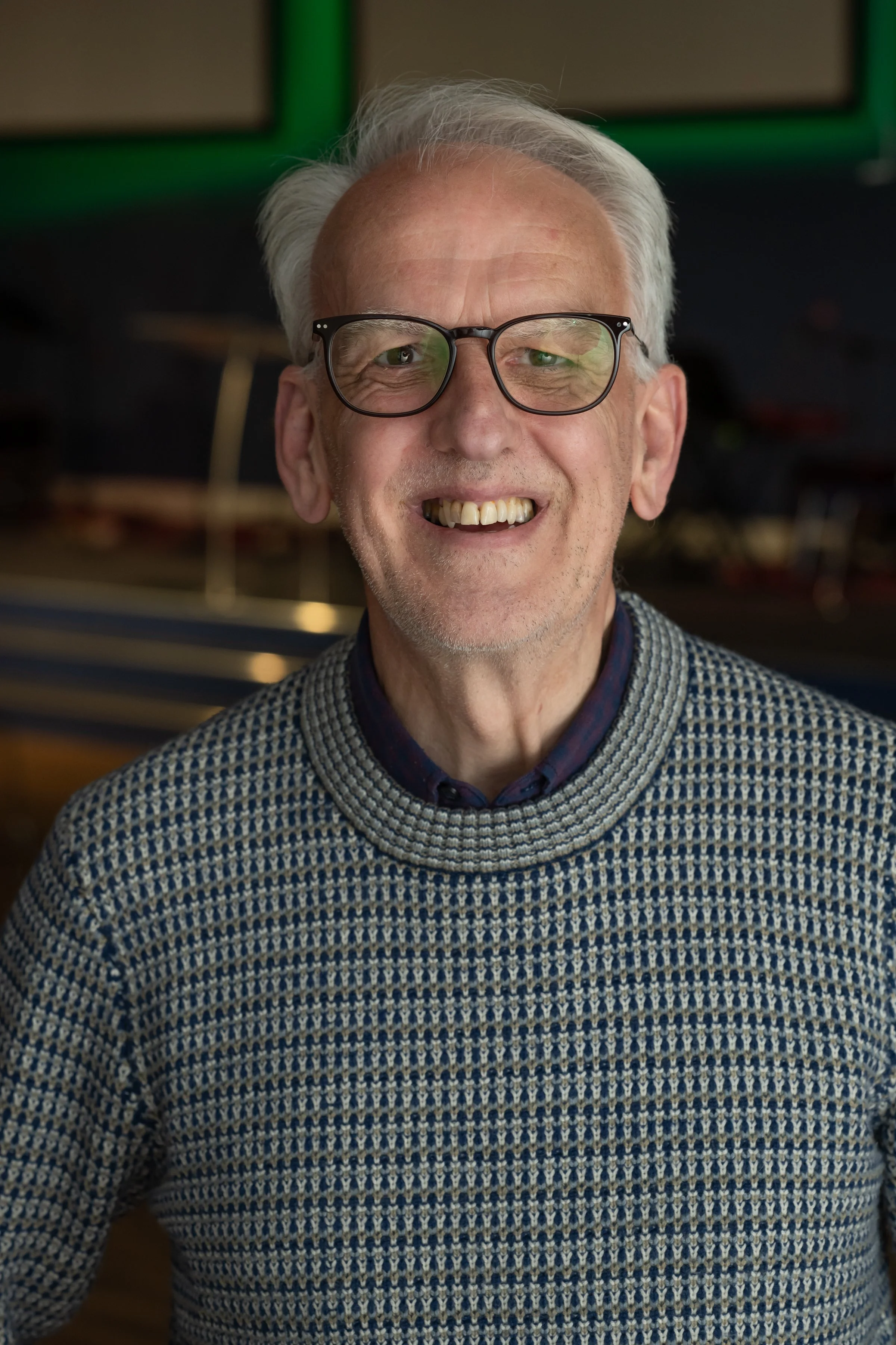 Close-up of an elderly man with white hair, glasses, smiling, wearing a patterned sweater and collared shirt, indoors with green and black background.