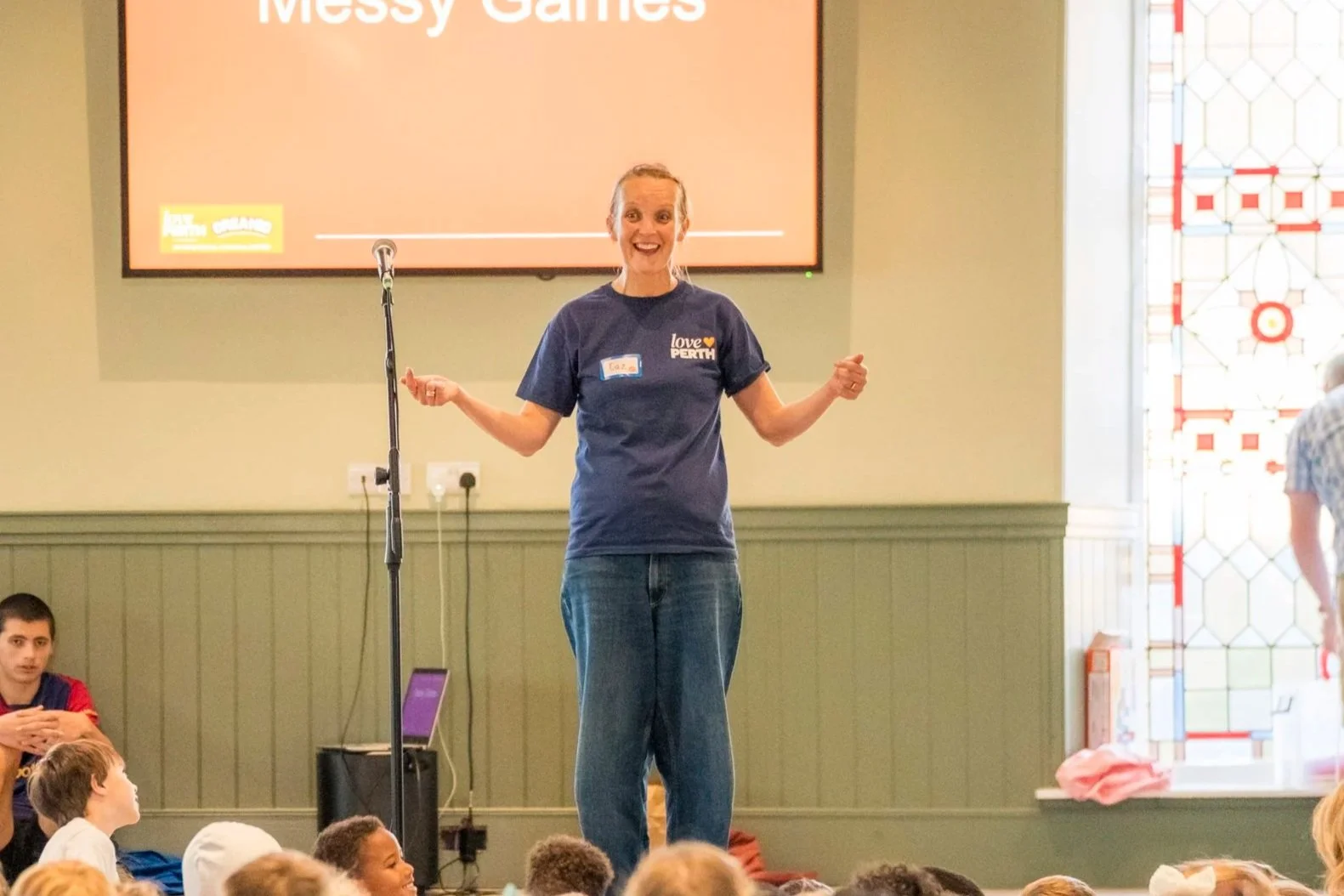 A woman in a blue t-shirt standing on a stage, smiling, with children sitting on the floor in front of her. A large screen behind her displays the words "Messy Games."