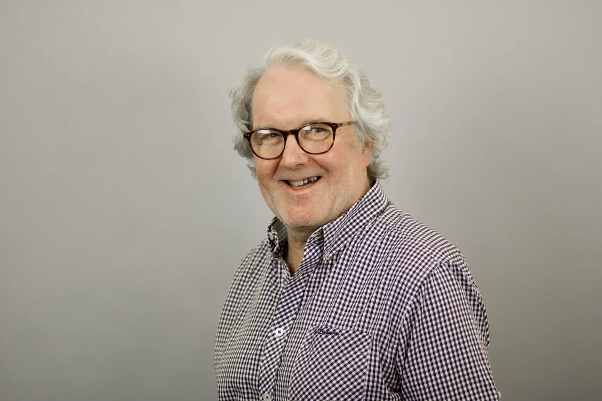 An older man with gray hair, glasses, and a checkered shirt smiling in front of a plain gray wall.