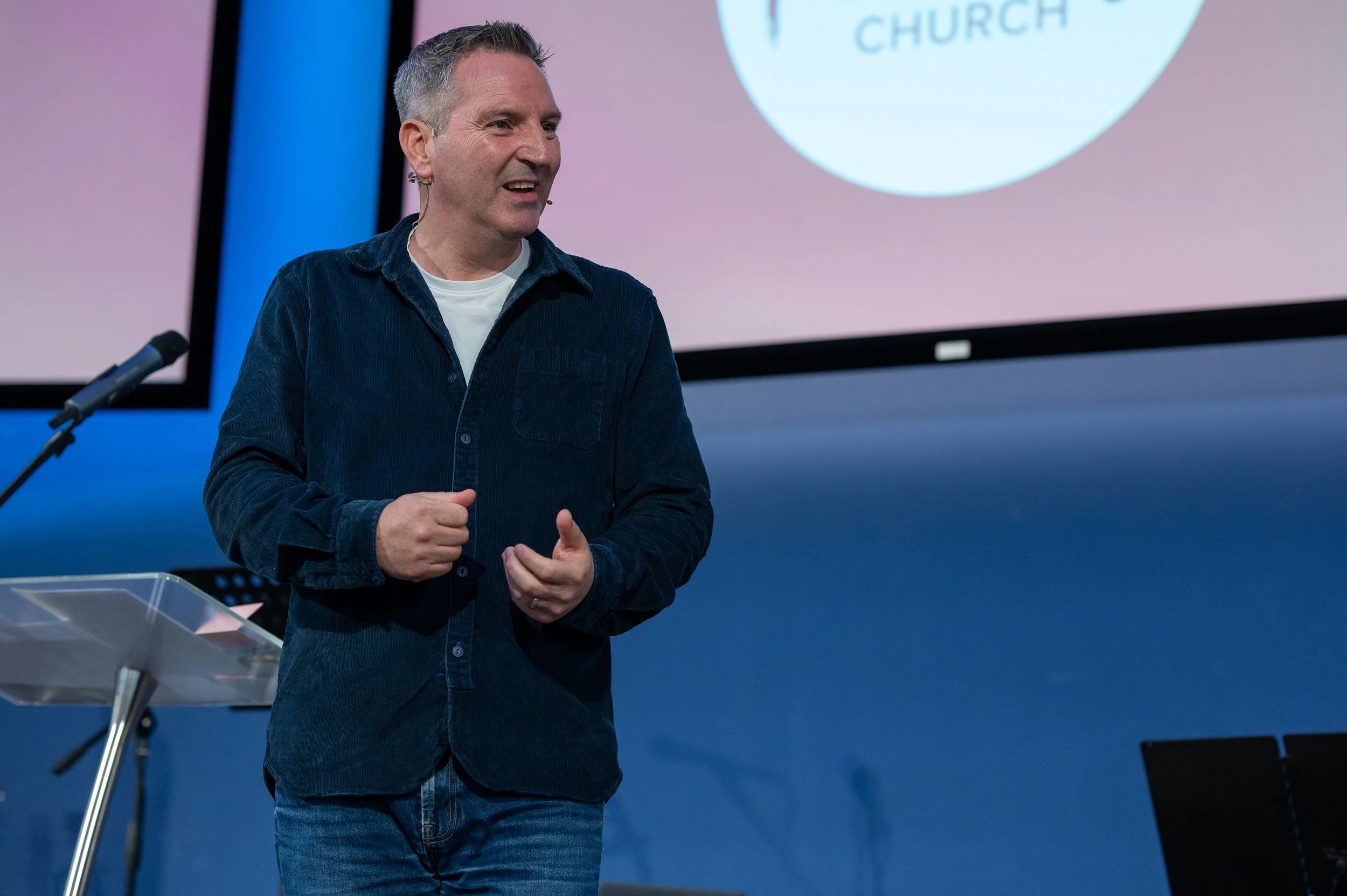 A man with gray hair and a earpiece speaking at a podium in front of a large screen displaying part of a logo that reads "CHURCH." He is wearing a dark blue corduroy shirt and jeans, and is gesturing with his hands.