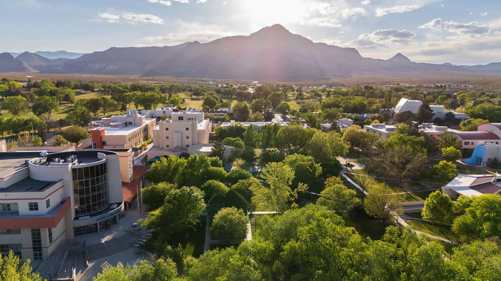 Aerial view of a university campus with modern buildings surrounded by green trees, with mountains in the background and a partly cloudy sky.