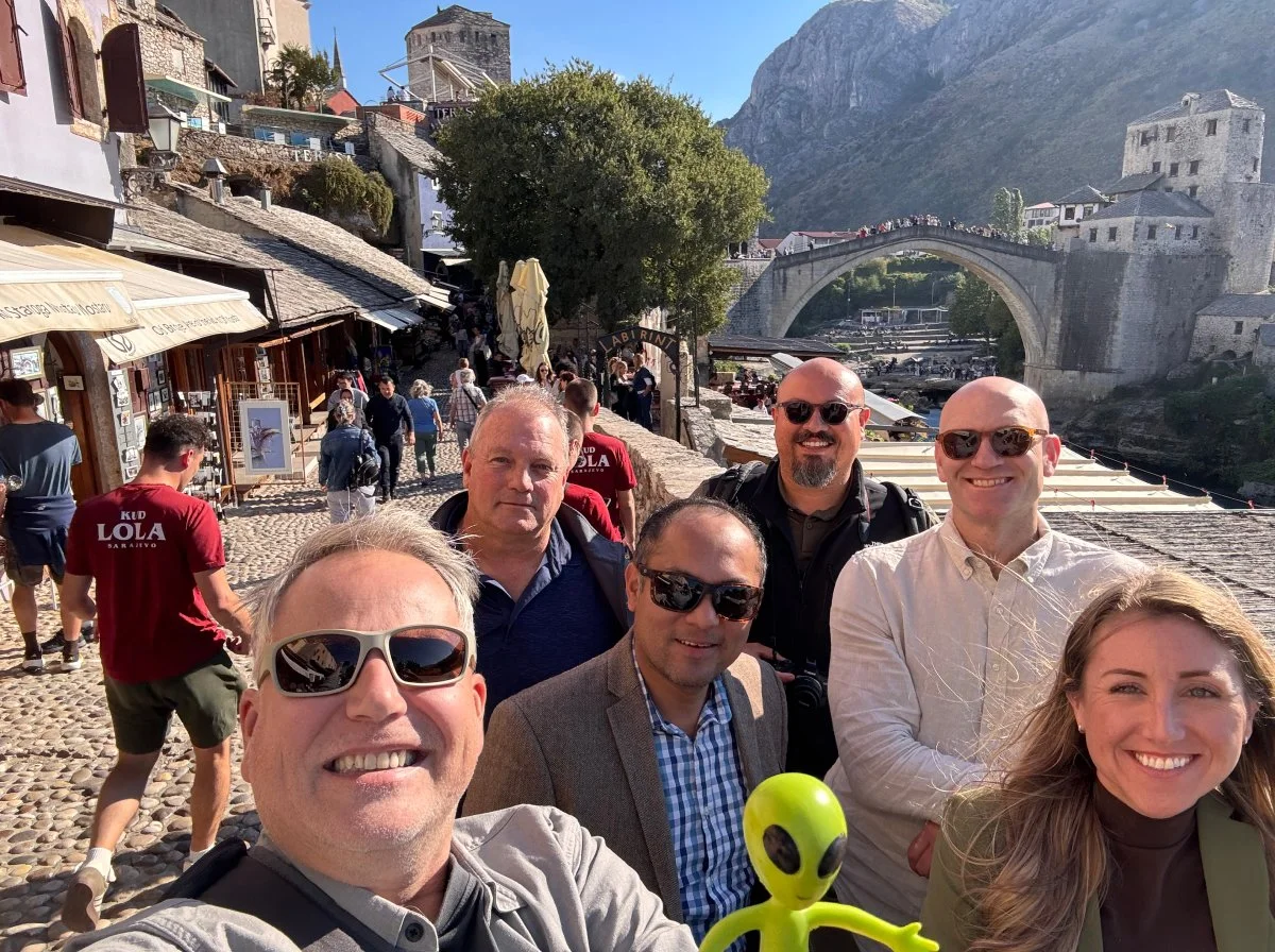 Group of six smiling people taking a selfie on a cobblestone street in a European town with a stone bridge, historic buildings, and mountains in the background.