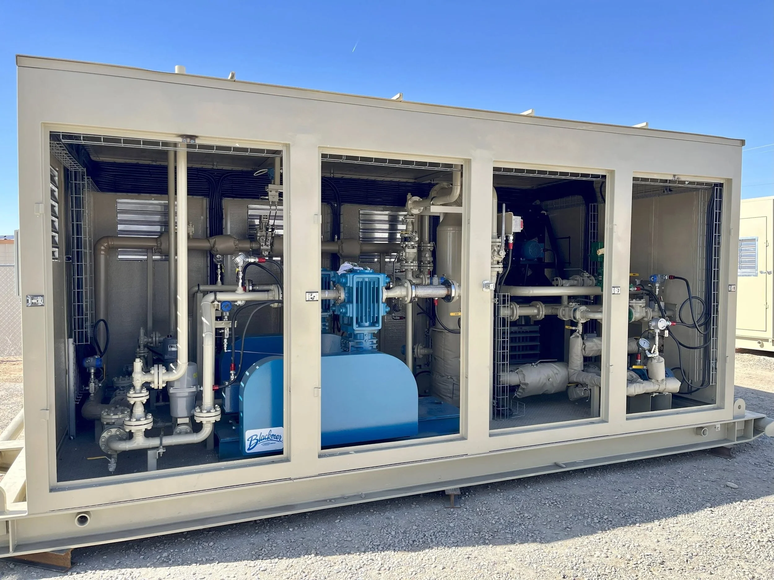 Industrial water treatment or pump system with pipes, valves, and gauges inside a metal enclosure, outdoor setting with clear blue sky.