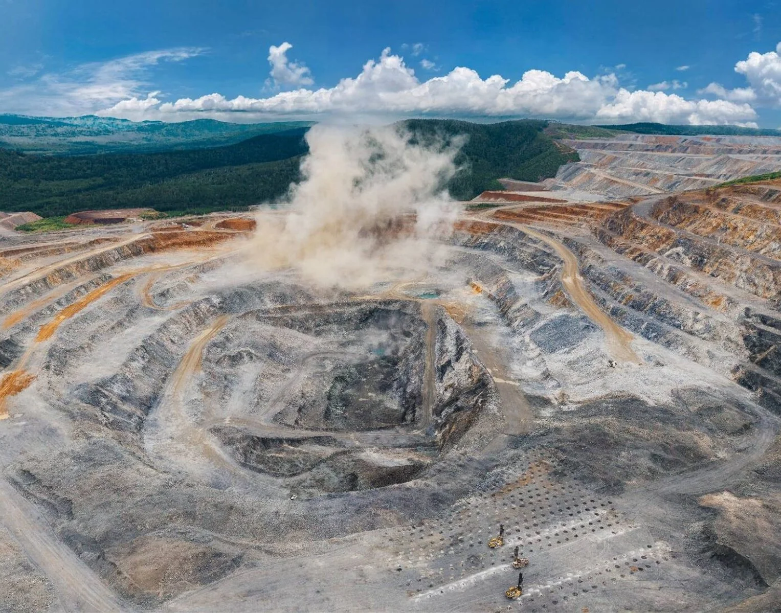 An open-pit mining operation with terraced excavations and a large plume of dust or steam rising from the central area, surrounded by mountainous terrain and a partly cloudy sky.