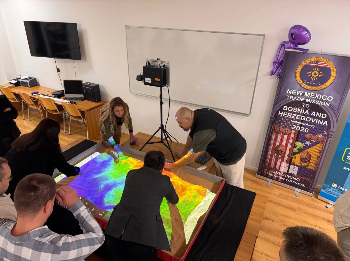 People in a classroom interacting with a large, colorful, interactive topographic map of a landmass, with a banner in the background titled 'New Mexico Trade Mission to Bosnia and Herzegovina 2026'.