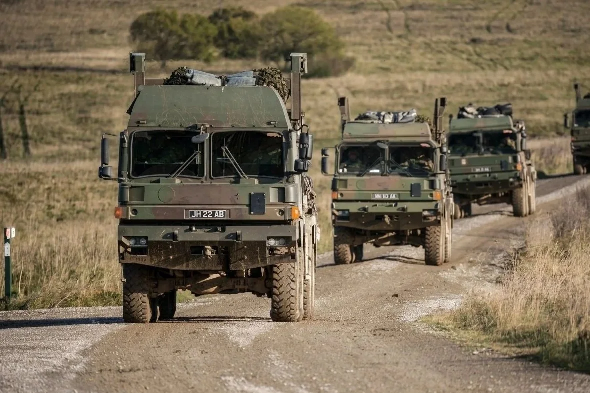 A convoy of military vehicles driving down a dirt road through a rural area with grassy fields and rolling hills.