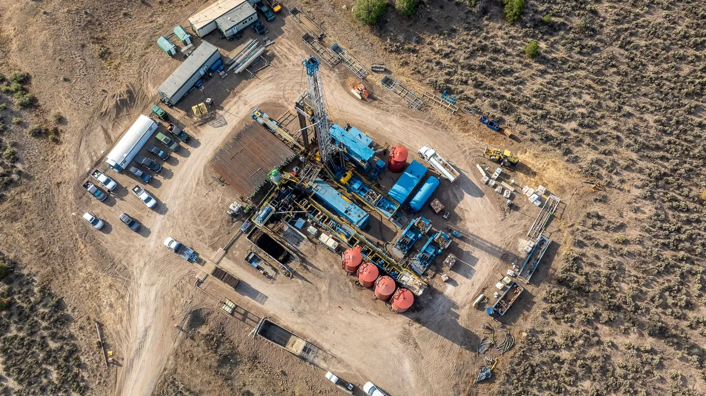 An aerial view of an oil drilling site in a desert area with multiple vehicles, equipment, and storage tanks surrounding the rig.
