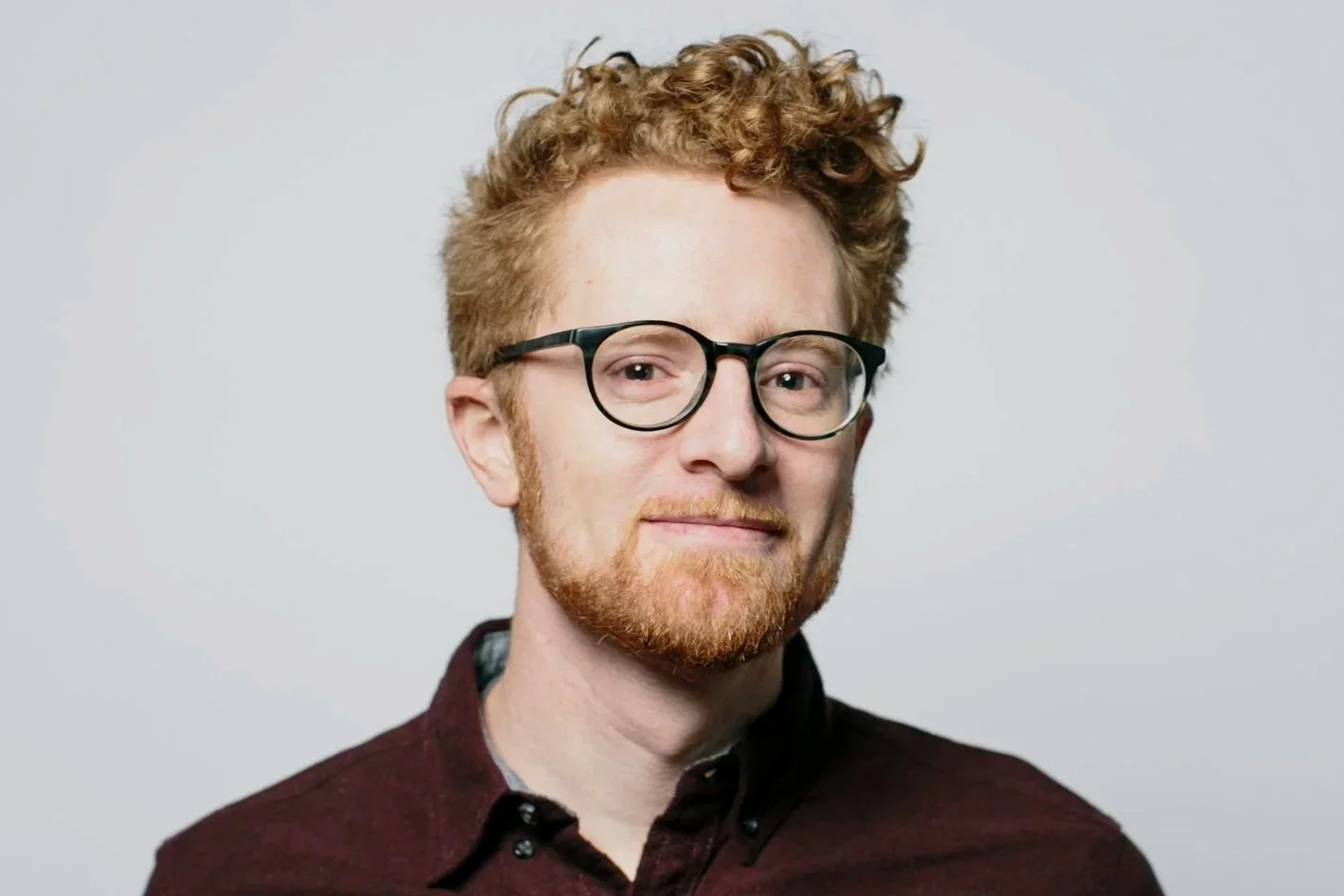 A man with curly red hair, glasses, and a beard, smiling slightly, wearing a dark maroon shirt, against a plain light background.