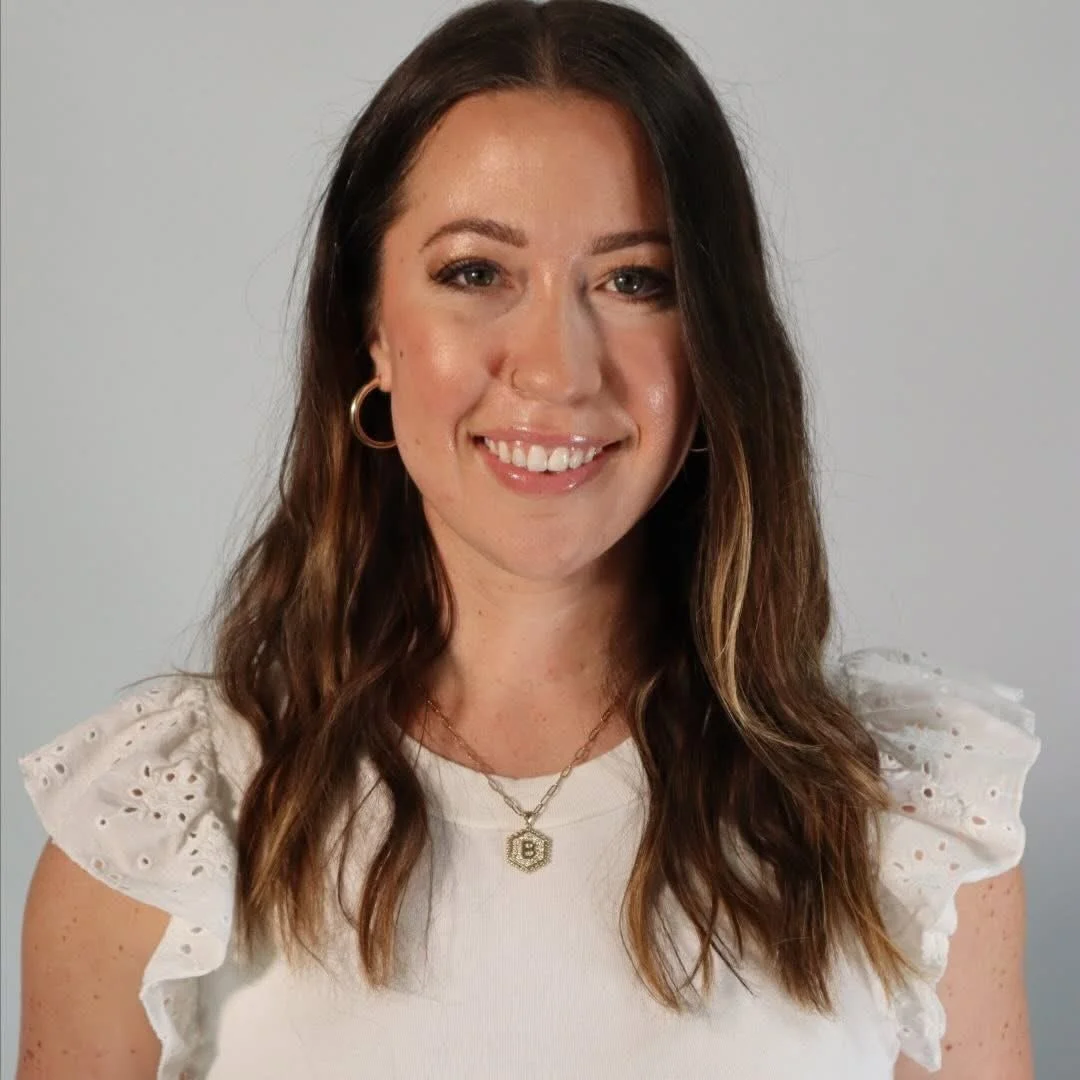 A woman with long, wavy brown hair and hoop earrings, smiling, wearing a white top with eyelet details on the sleeves, a gold necklace with a pendant, and a smaller chain with a initial pendant.