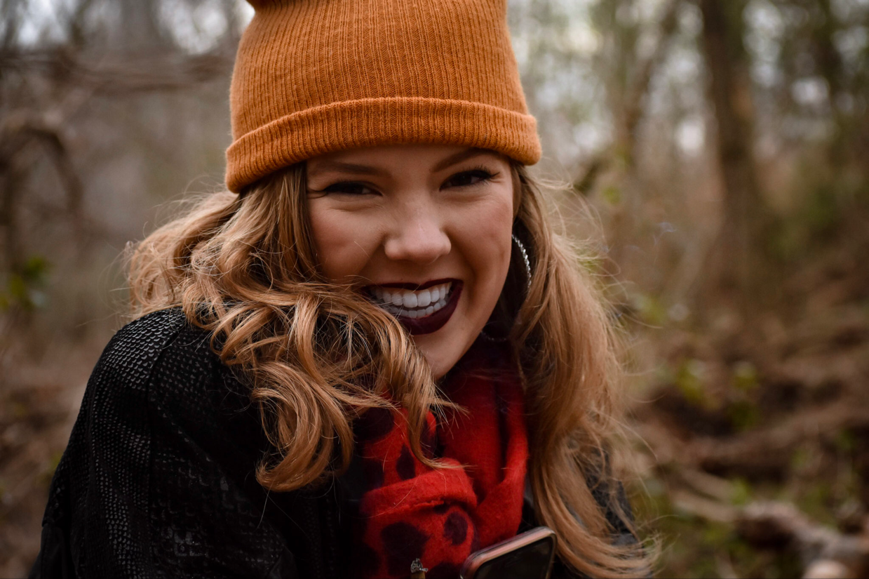 A young woman with wavy red hair wearing an orange knit beanie, dark lipstick, and a black jacket with a red scarf, smiling outdoors in a wooded area.