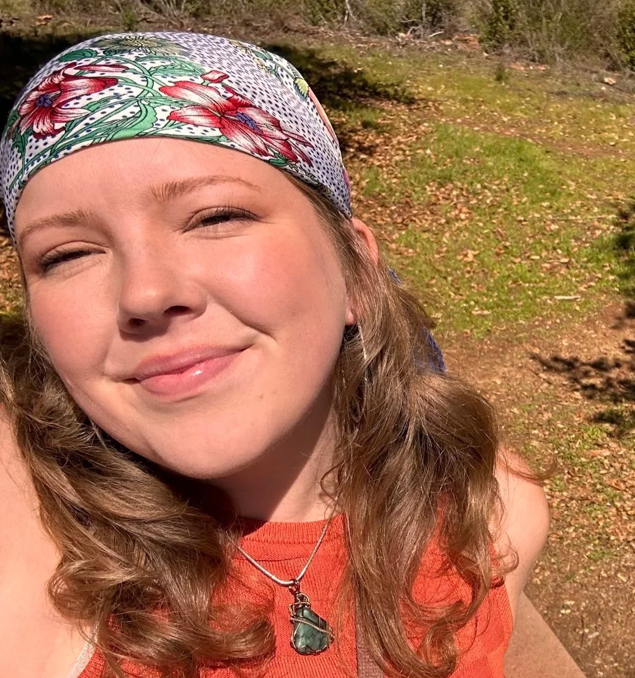 A young woman smiling outdoors with a colorful floral bandana on her head and an orange sleeveless top. She is wearing a pendant necklace with a green stone. The background shows grass and dirt with some sunlight.