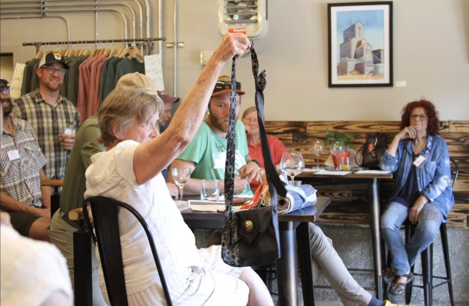 A group of people at a meeting or workshop, with one woman holding up a black and gray patterned dog leash or strap, seated at tables with glasses and papers, in a room with framed artwork and clothing racks in the background.