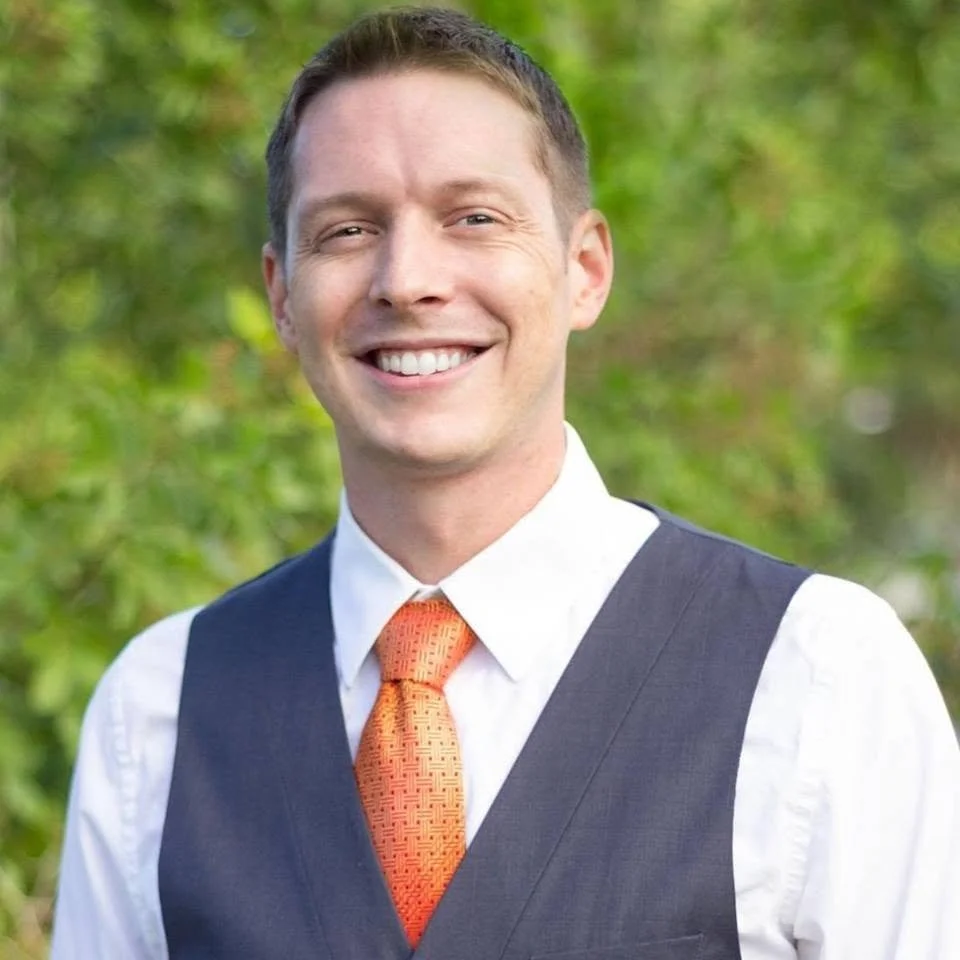 A smiling man in a white shirt, dark vest, and orange tie, standing outdoors with green foliage in the background.