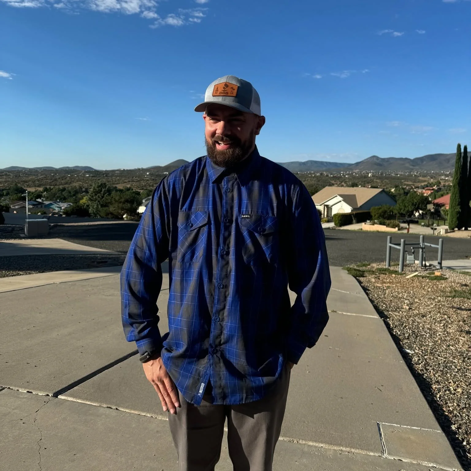 A man with a beard and mustache, wearing a gray and mesh baseball cap and a blue plaid shirt, standing outdoors on a sunny day with hills and houses in the background.