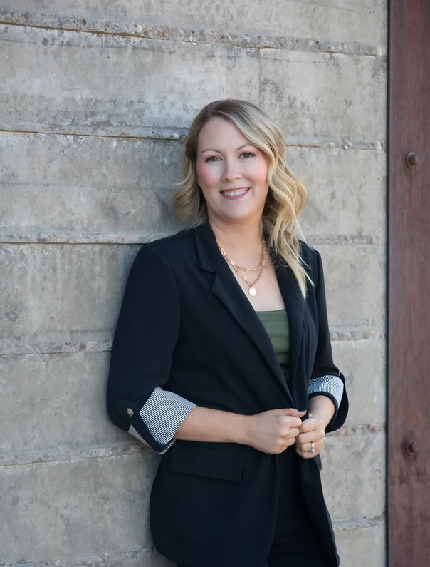 A woman with blonde wavy hair smiling, wearing a black blazer with striped cuffs, green top, and gold jewelry, standing against a concrete wall.