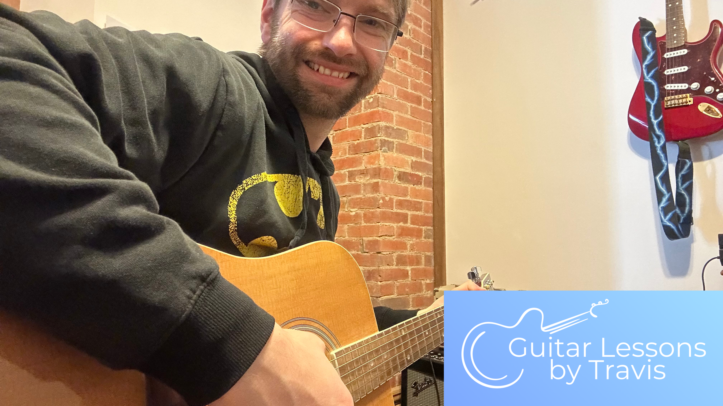 A man with glasses, facial hair, and a black hoodie with yellow design, smiling and playing an acoustic guitar indoors with a brick wall and a red electric guitar on the wall in the background.