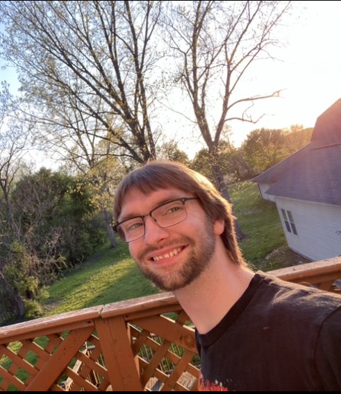 A young man with glasses and a beard smiling outdoors on a wooden deck during sunset, with trees and a white house in the background.