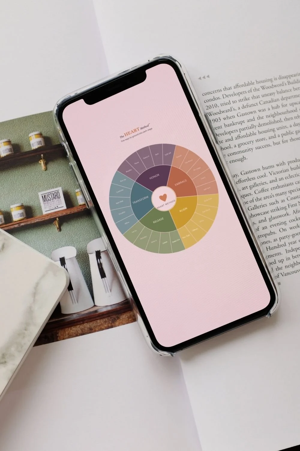 Smartphone displaying a circular diagram titled 'The HEART Method' with sections labeled 'Embrace,' 'Align,' 'Release,' 'Transform,' 'Honor.' The phone is placed on an open book with text and is partly on a white surface, with a blurred background of mustard jars on a green wall shelf and kitchen items.