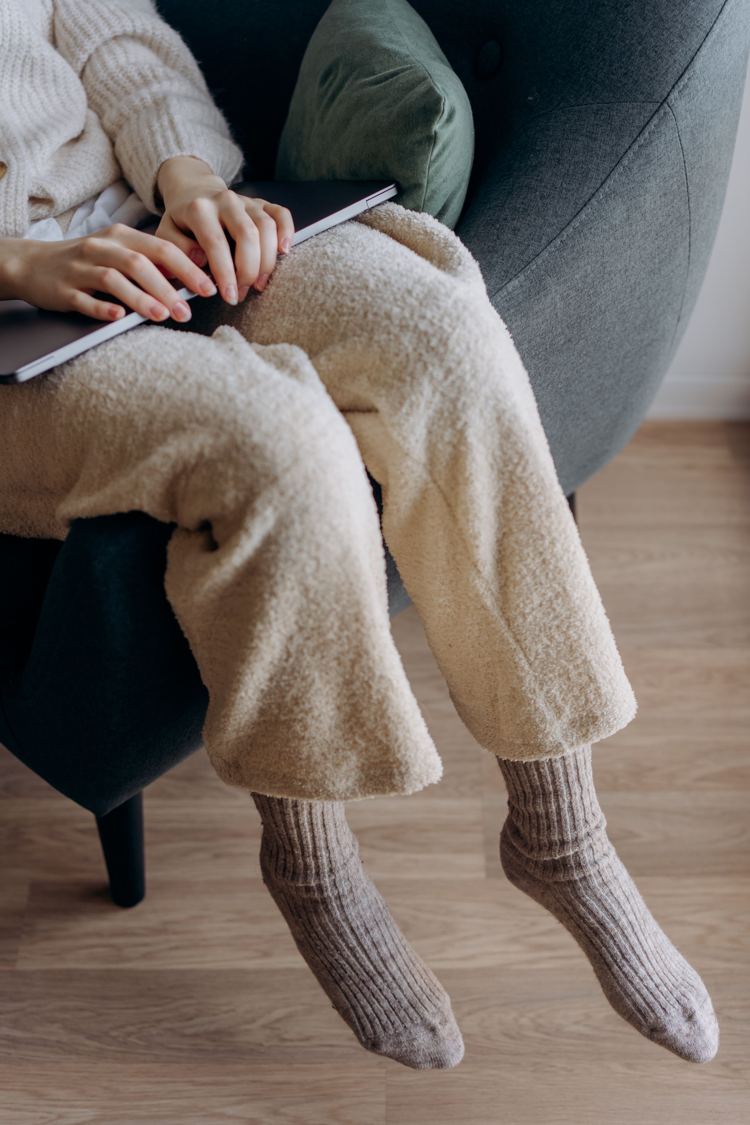 A person sitting on a gray upholstered chair wearing thick, beige, striped socks and beige fuzzy pants. Their black tablet or laptop rests on their lap, and they are wearing a cream-colored sweater and green pants.