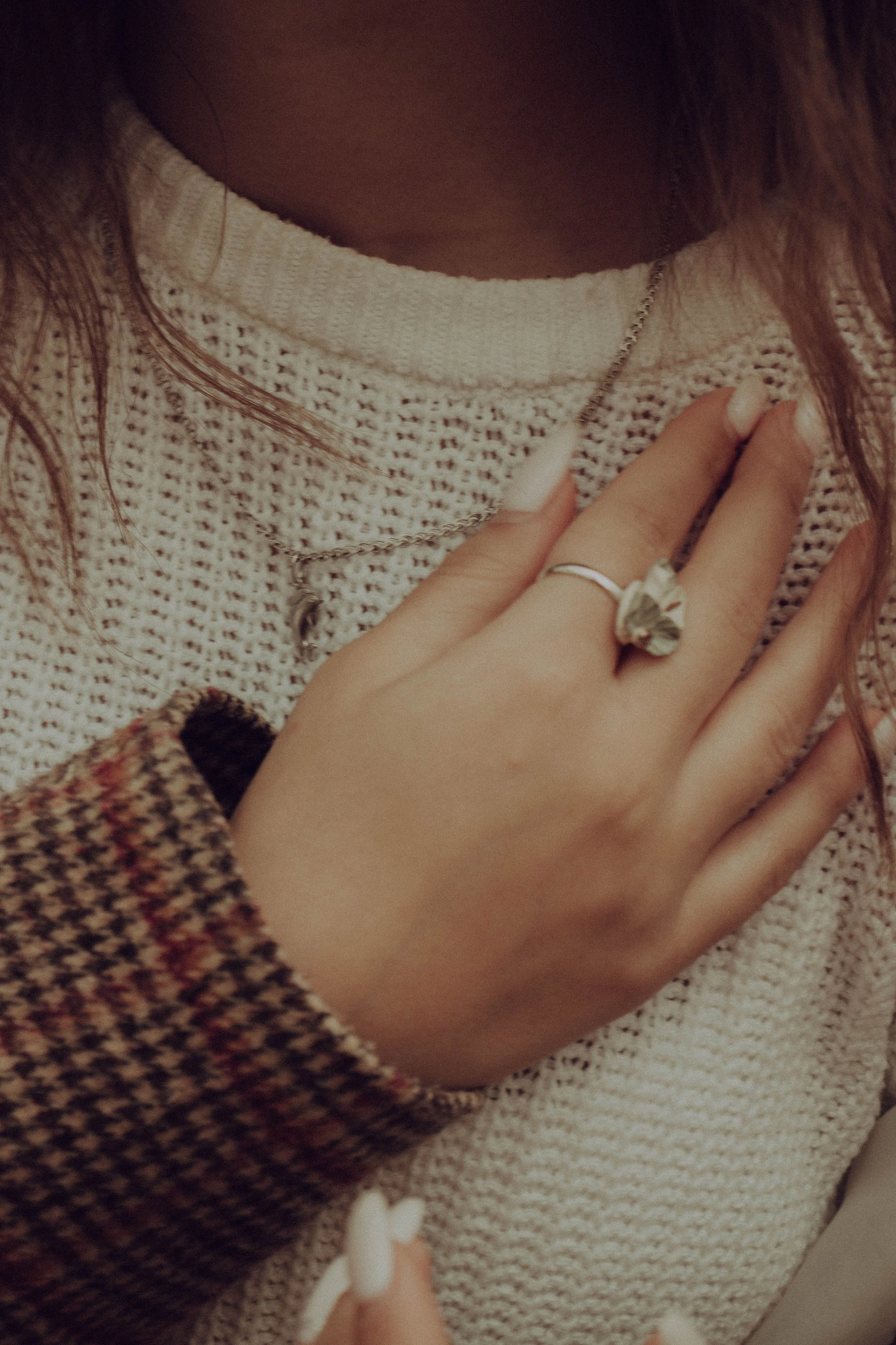 Close-up of a person's hand with a ring on the finger, touching their chest which is clothed in a beige, patterned sweater. The person's face is not visible.