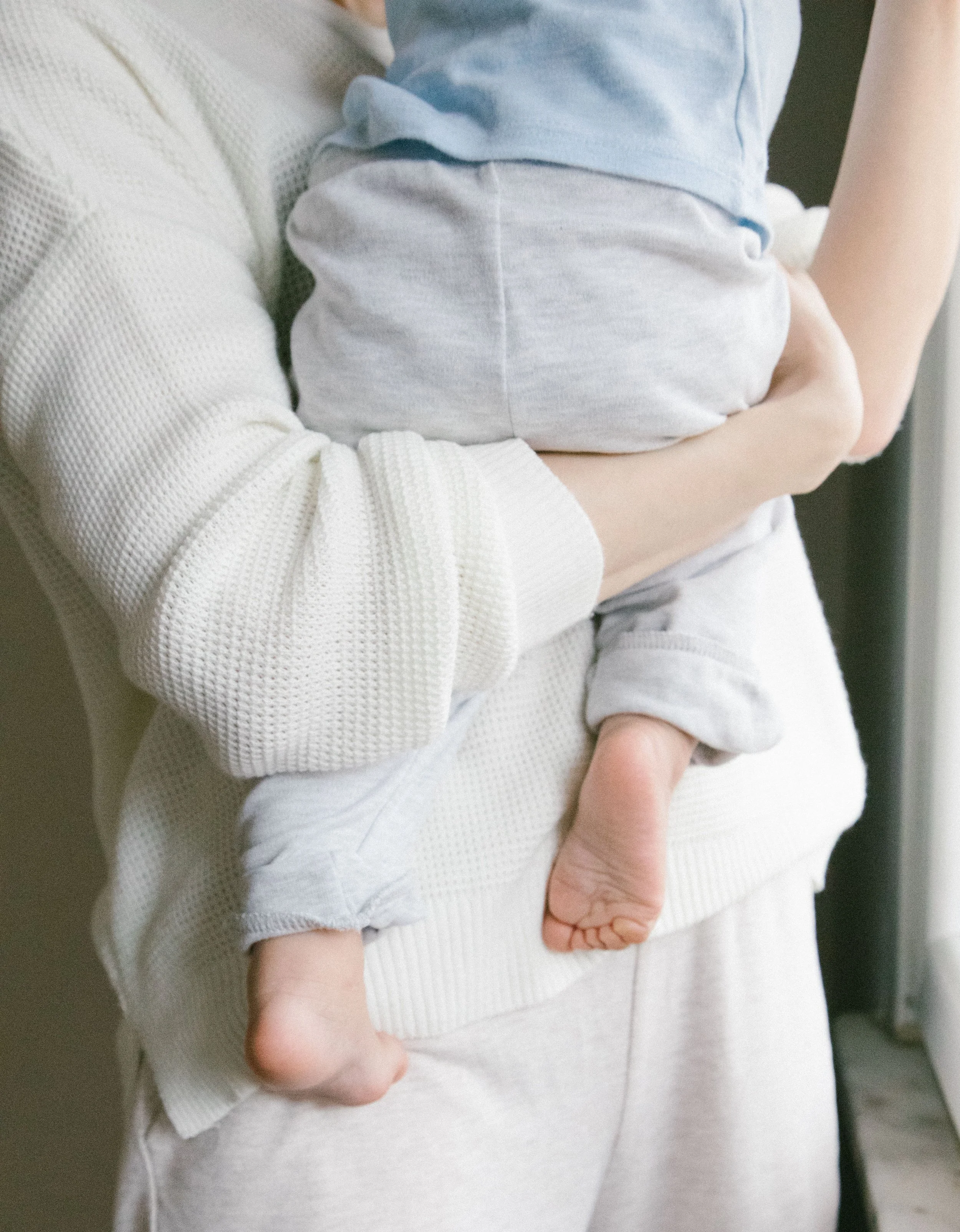 A person holding a baby, with the focus on the baby's hand and the person's arm. The baby is dressed in a light-colored outfit, and the person is wearing a white textured sweater.