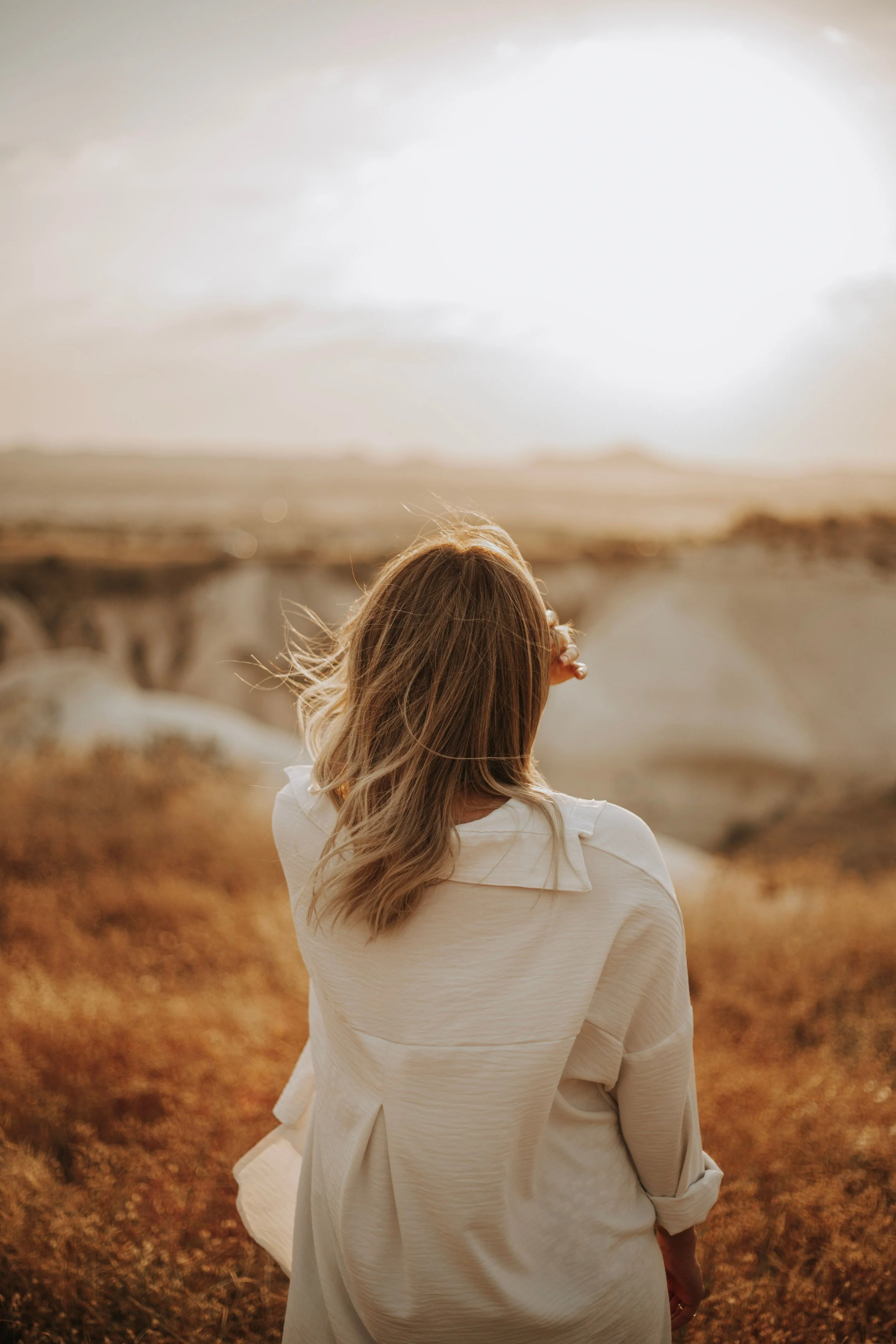 A woman with long, wavy hair wearing a cream-colored dress standing in a field during sunset or sunrise. soul searching