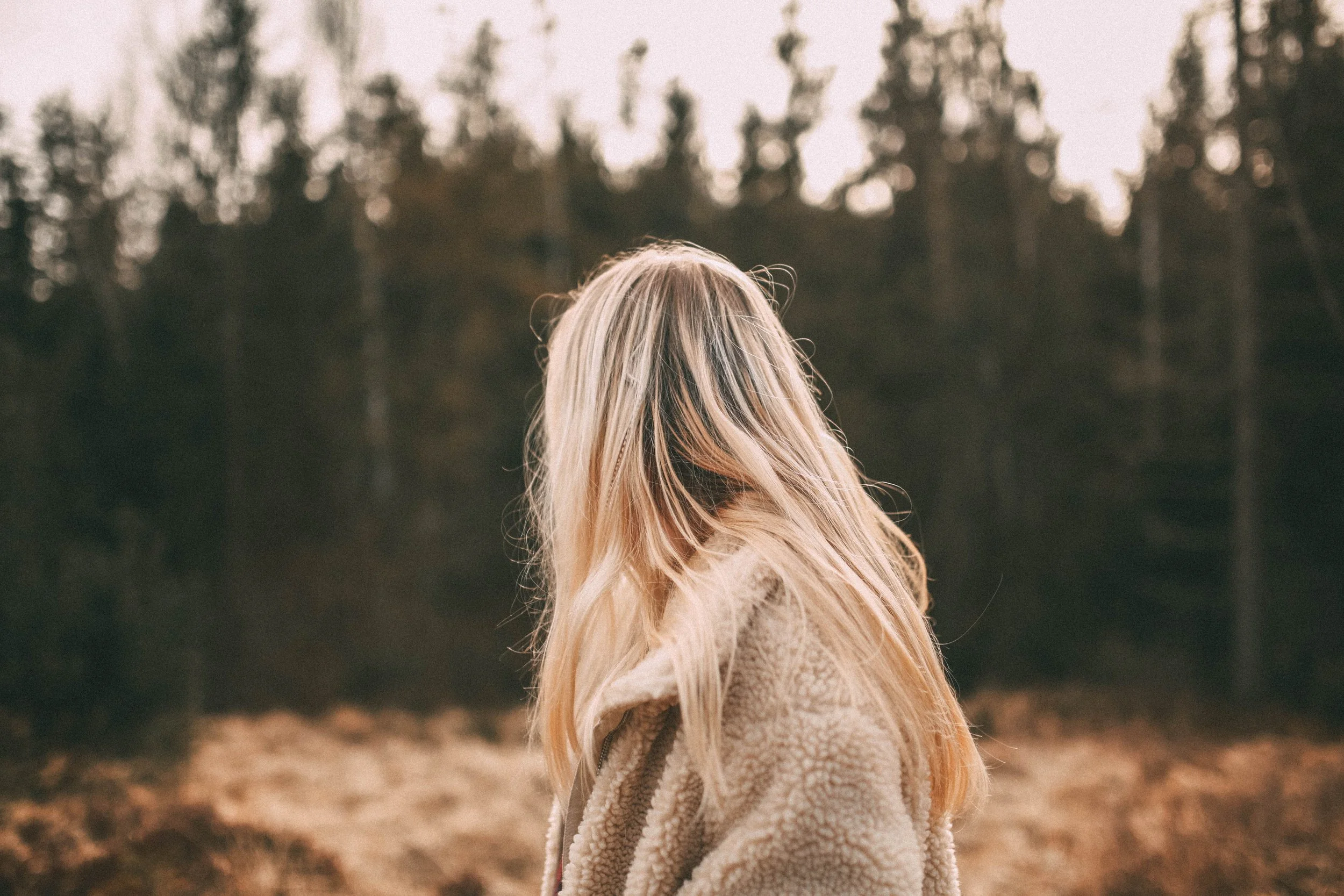 A woman with long blonde hair standing outdoors in a forest, facing away from the camera.