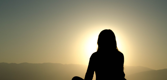 Silhouette of a woman sitting outdoors during sunset or sunrise with mountains in the background.