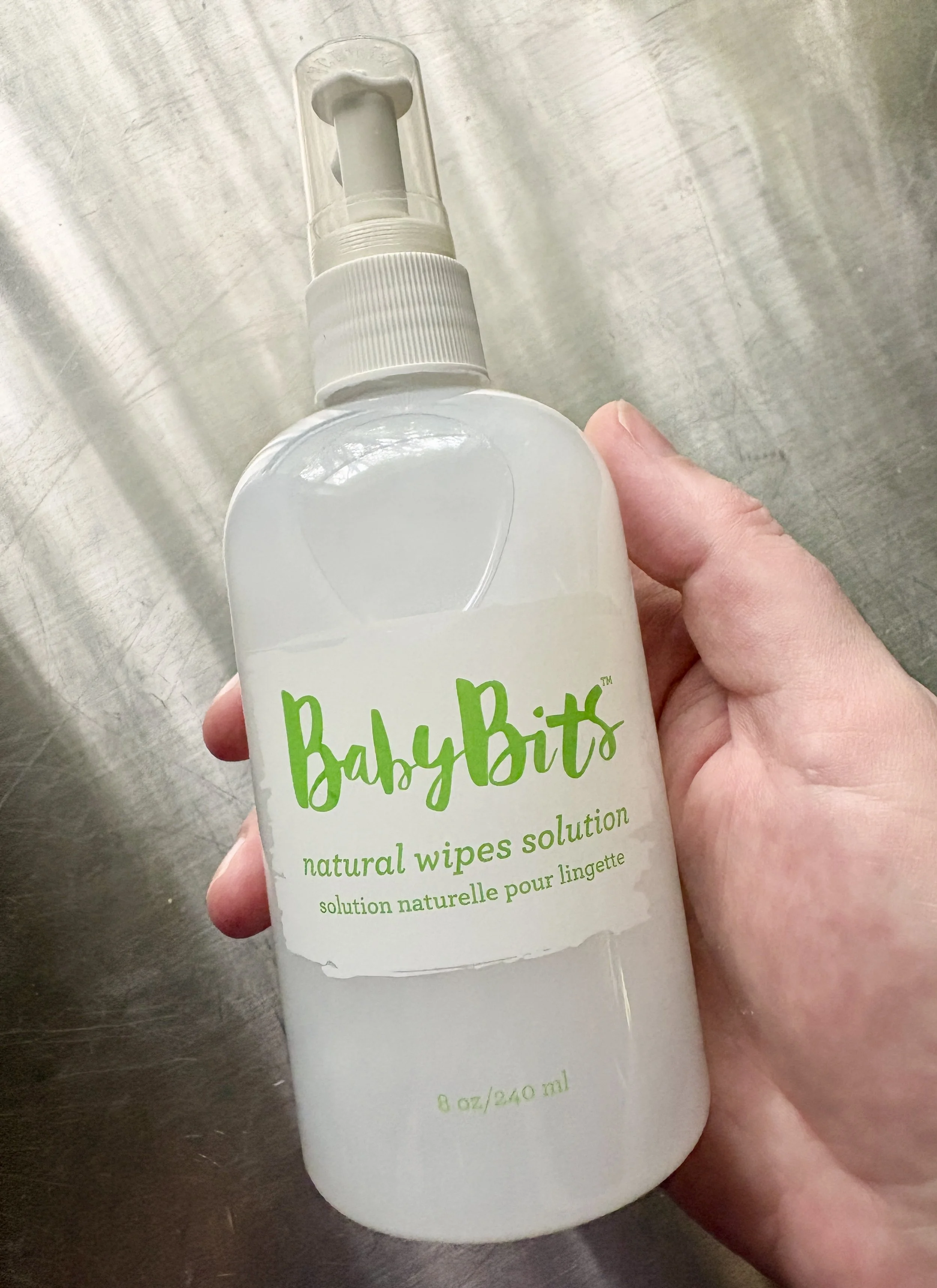 A person holding a white spray bottle of Baby Bits natural wipes solution on a metallic surface.