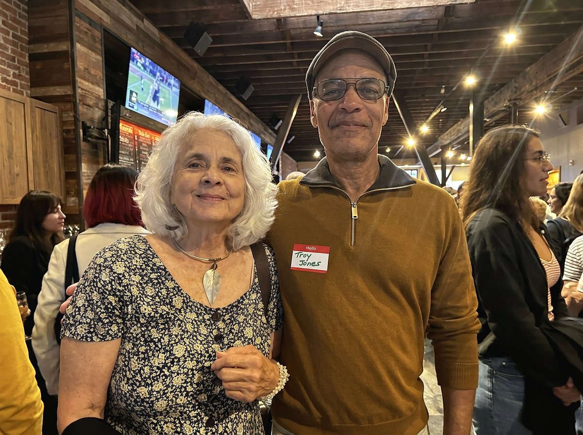 An elderly woman with white hair wearing a patterned dress and a large leaf-shaped necklace, standing next to a middle-aged man in a brown sweater and glasses, in a bustling indoor setting with many people, TV screens, and warm lighting.